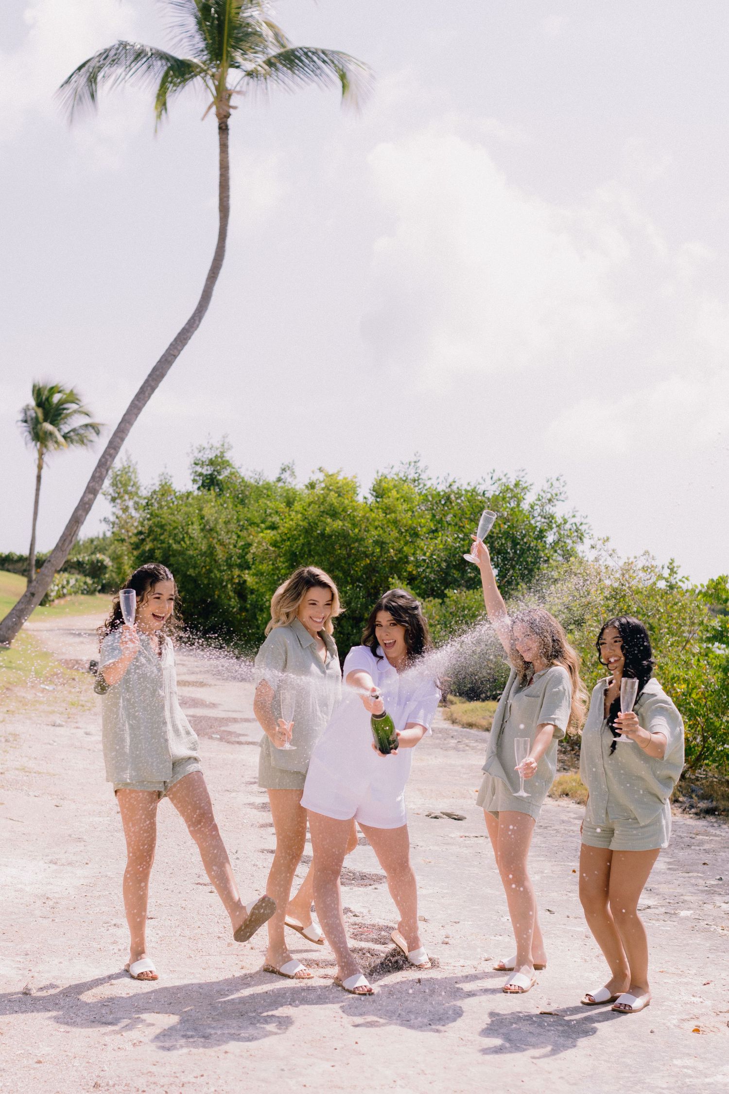 Five women celebrating on a tropical beach path, popping champagne bottle, wearing matching sage green outfits.