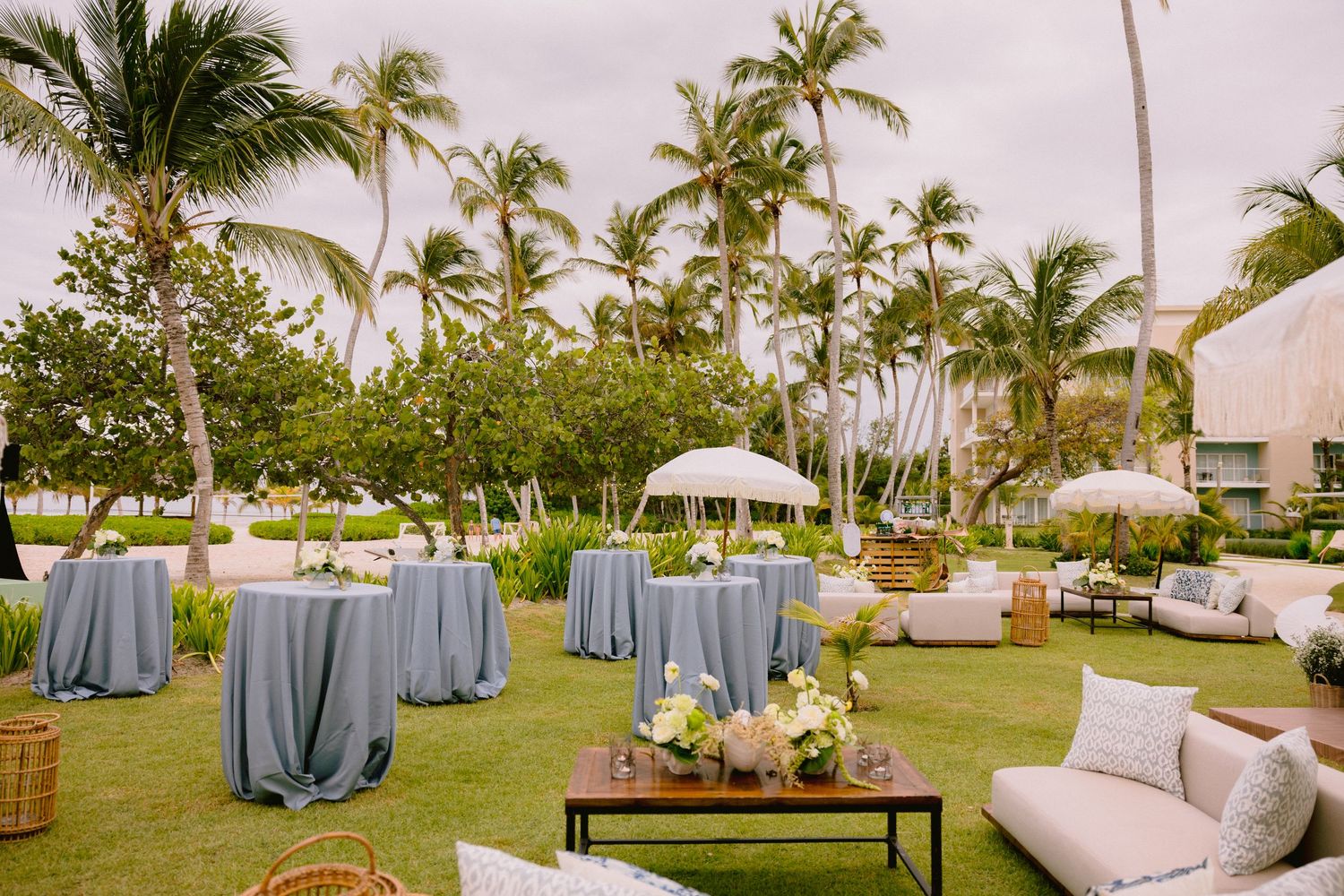 Outdoor tropical resort event space with blue-draped cocktail tables, palm trees, white umbrellas and lounge seating on a manicured lawn.