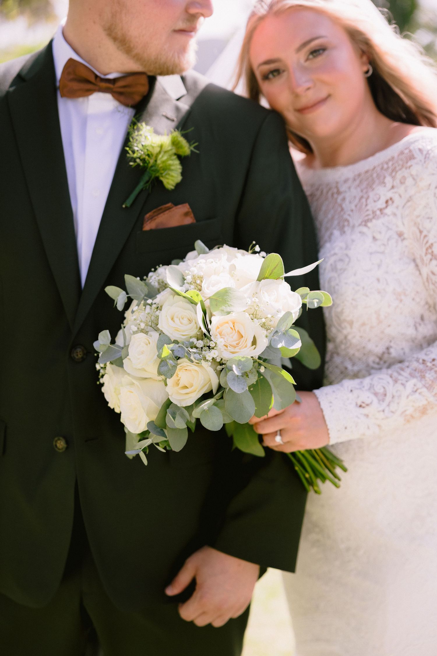 Bride in lace dress holds white rose bouquet beside groom in black suit with brown bow tie on wedding day.