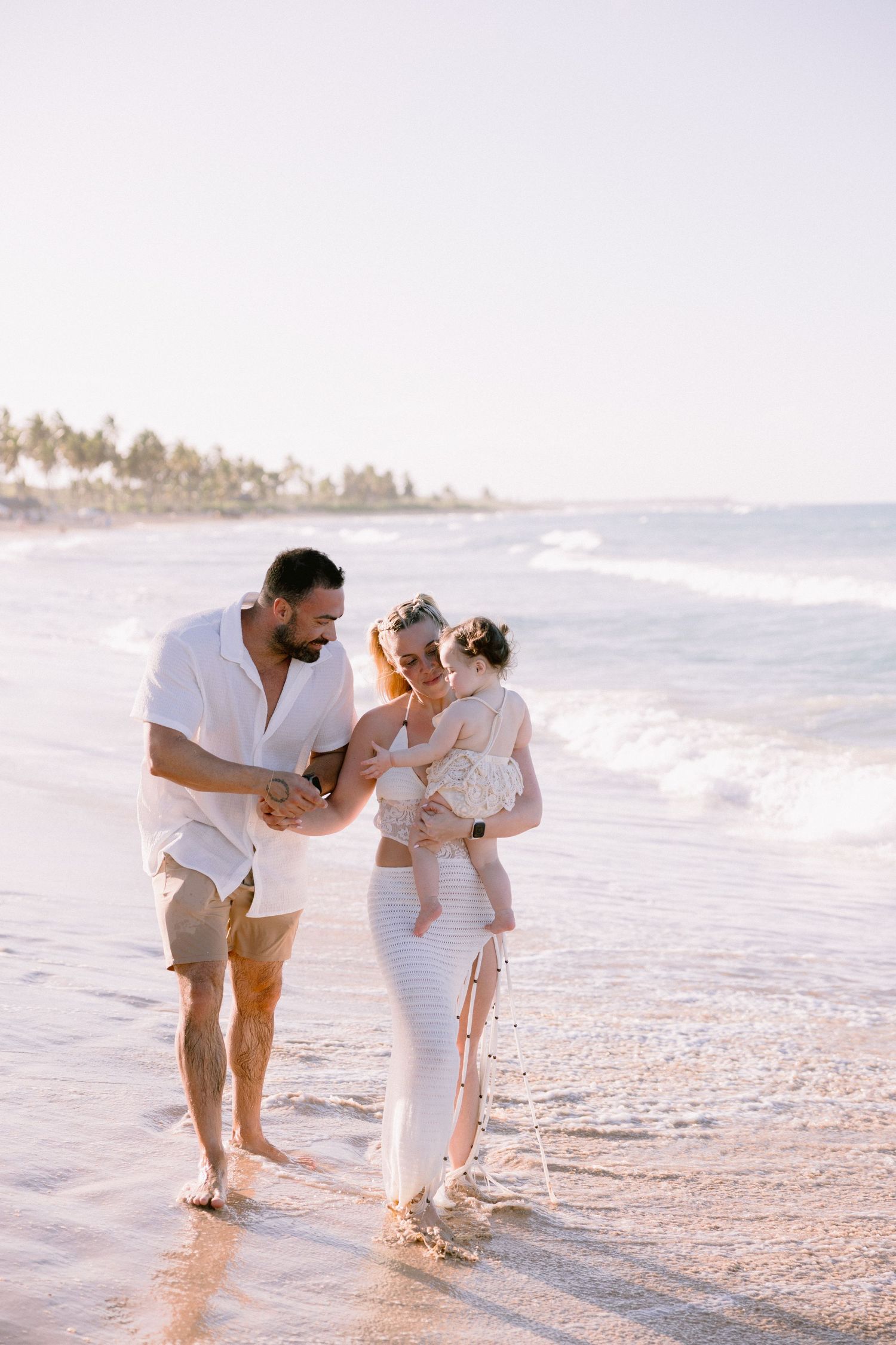 A happy family of three walks along a sunny tropical beach, with parents admiring their baby girl.