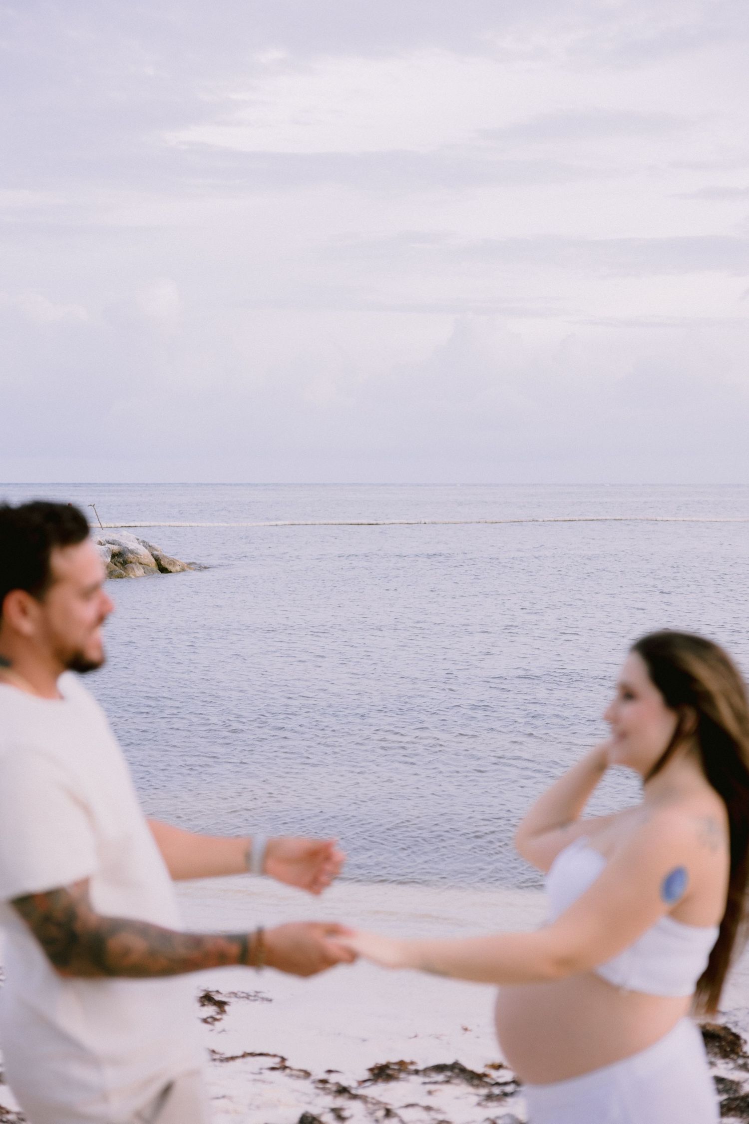 A couple in beach attire dances together on a white sand beach against an overcast sky.
