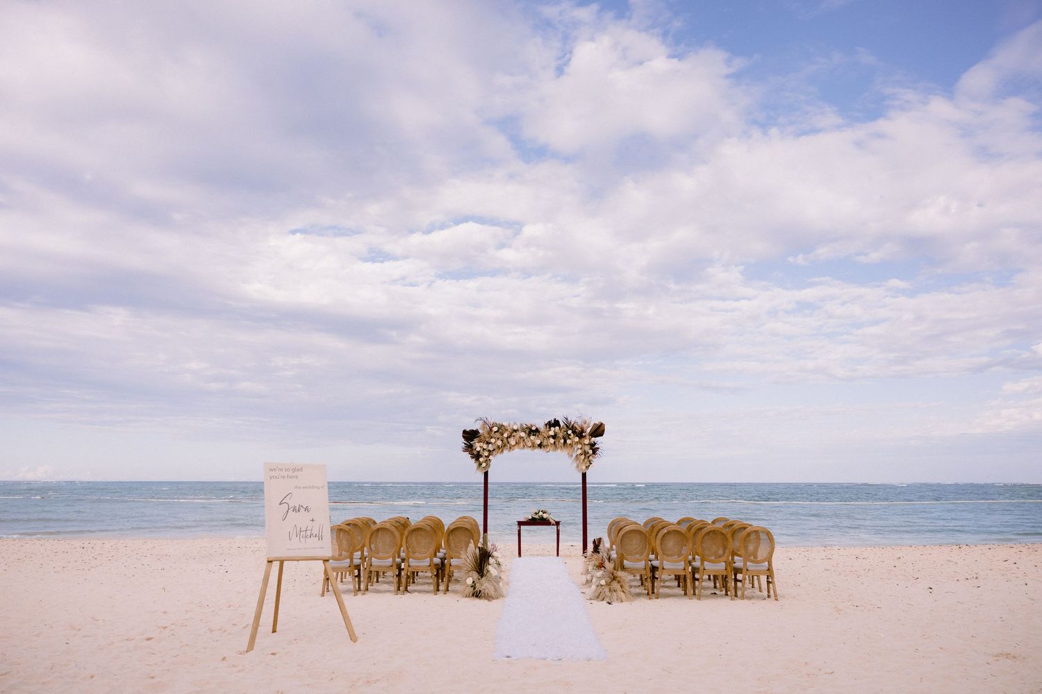 Elegant beach wedding ceremony setup with floral arch, wooden chairs, and white aisle on sandy shore under cloudy sky.