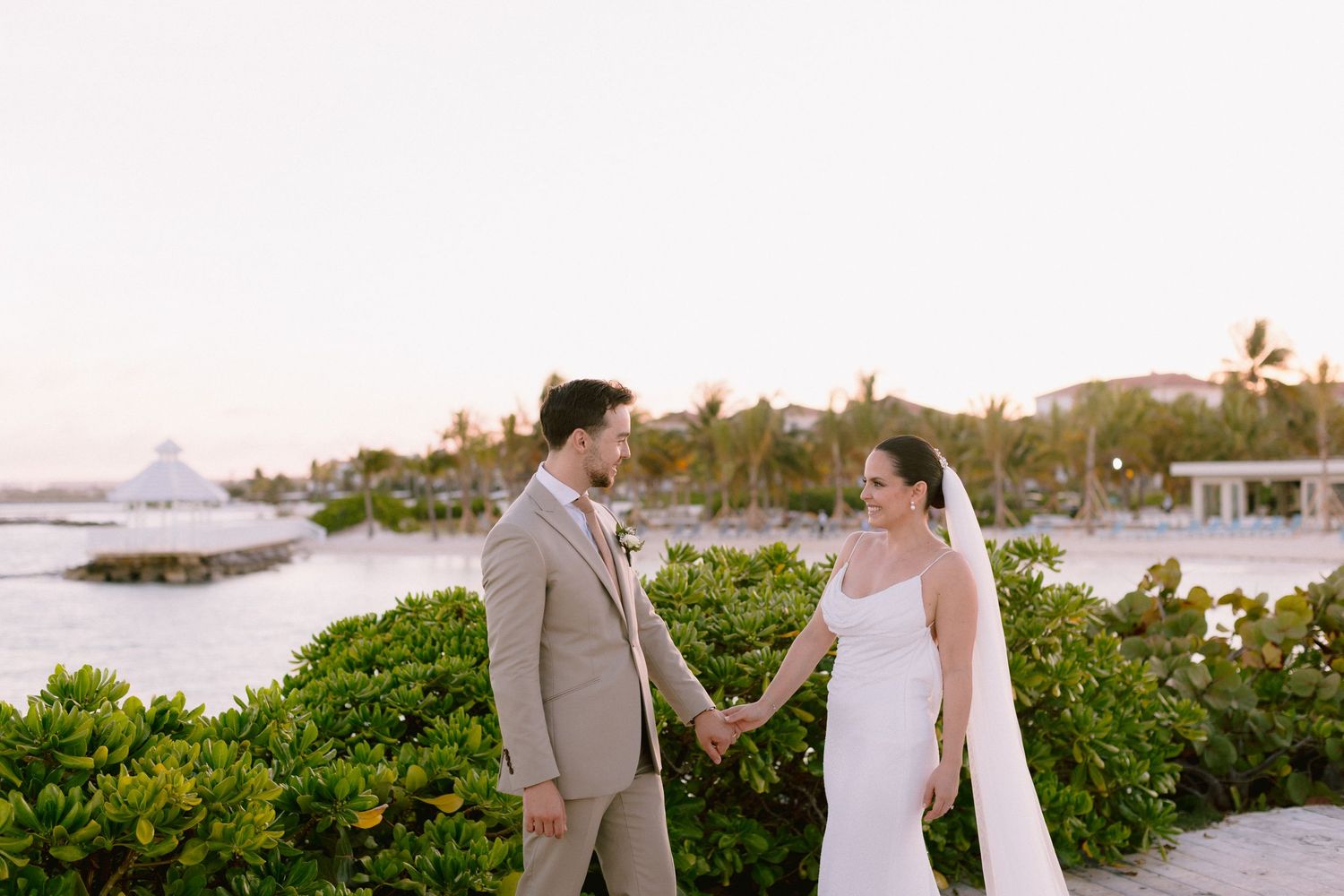 Bride and groom hold hands at sunset near tropical waterfront, bride in white gown with veil, groom in tan suit.