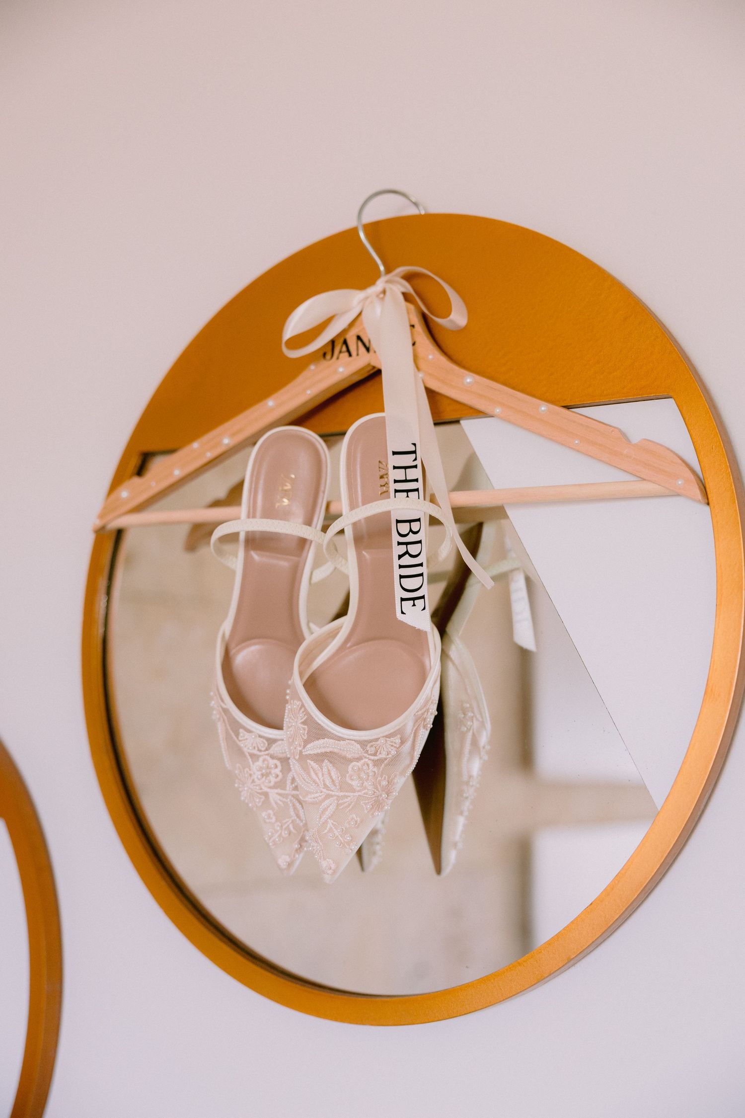 Elegant white lace bridal heels hanging on a wooden hanger labeled The Bride against a gold round mirror.