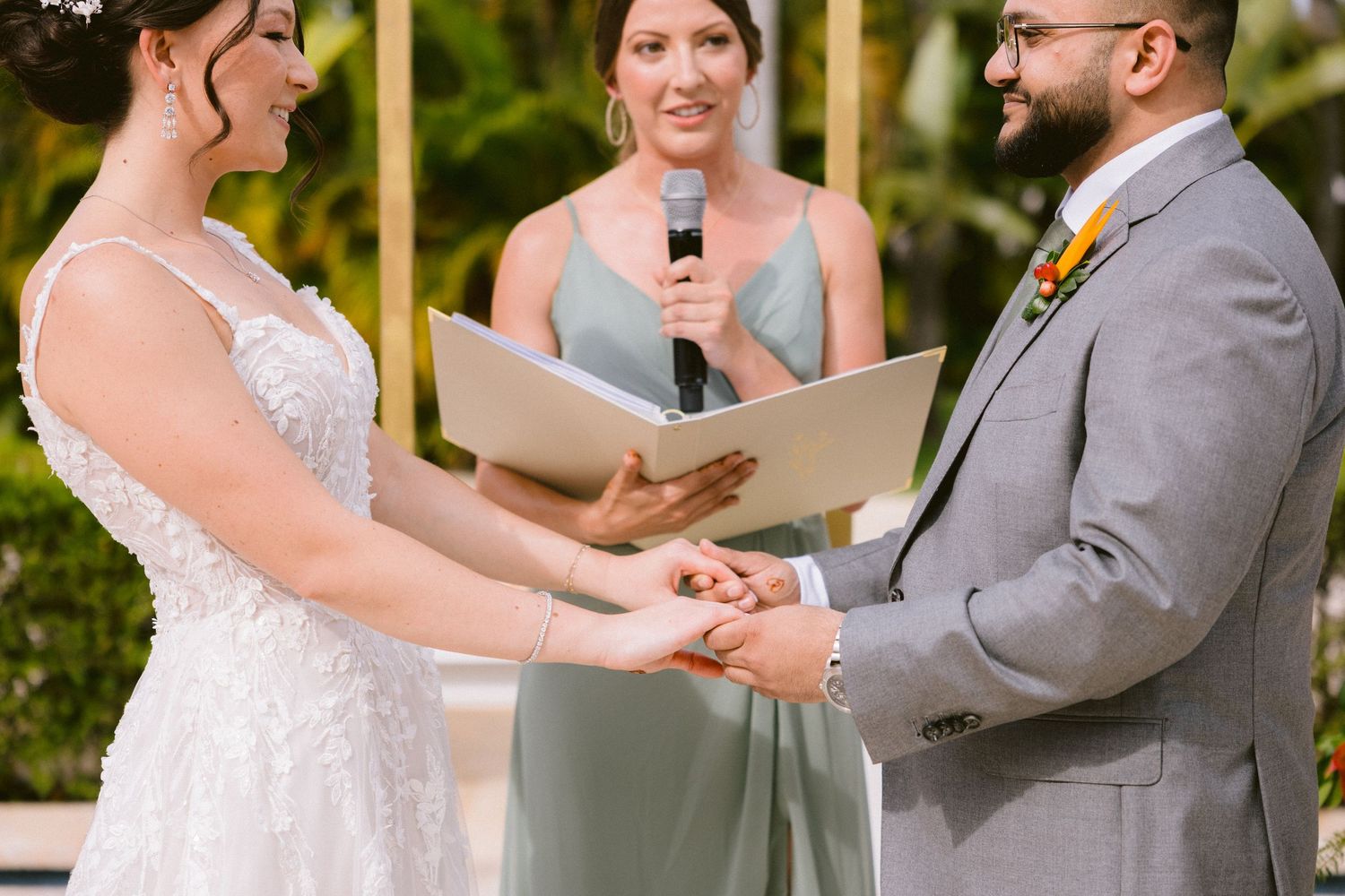 A couple holds hands during an outdoor wedding ceremony with lush greenery in the background.
