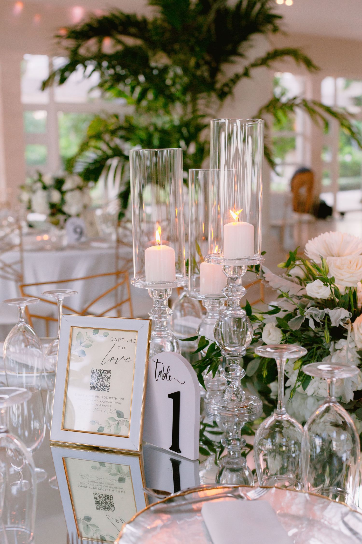 Elegant wedding table decor with glass candle holders, greenery garland, and table number in gold frame.