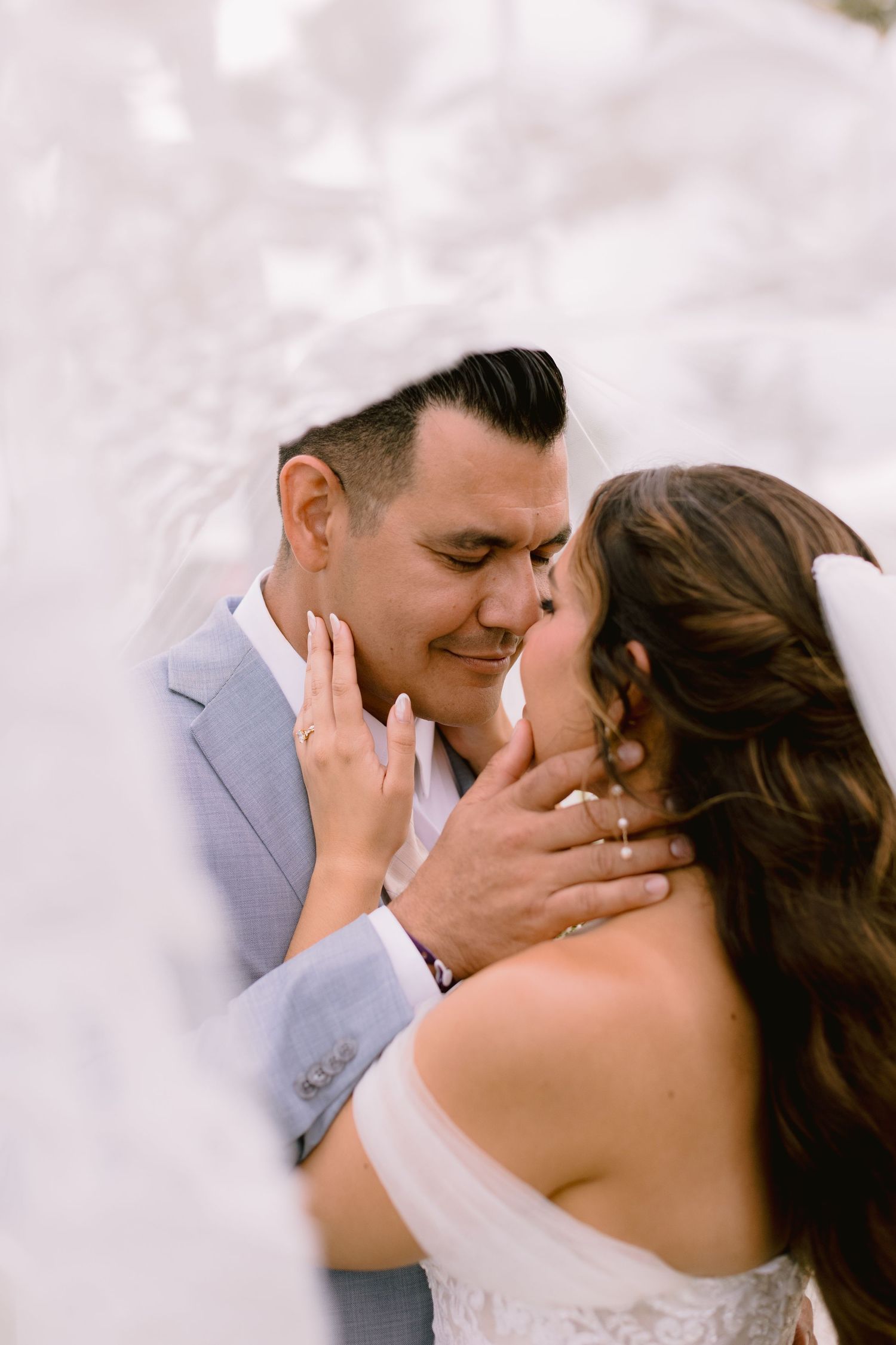 Bride and groom share a tender almost-kiss under a flowing veil on their wedding day.