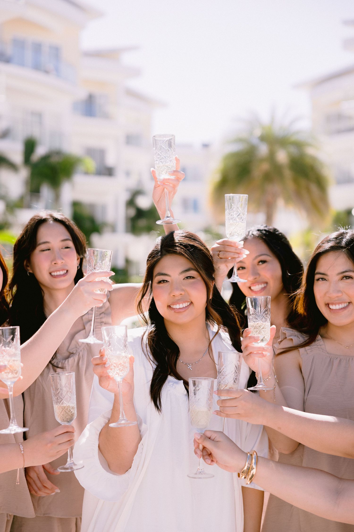 Bride and bridesmaids in neutral dresses raising champagne glasses in a celebratory toast outdoors.