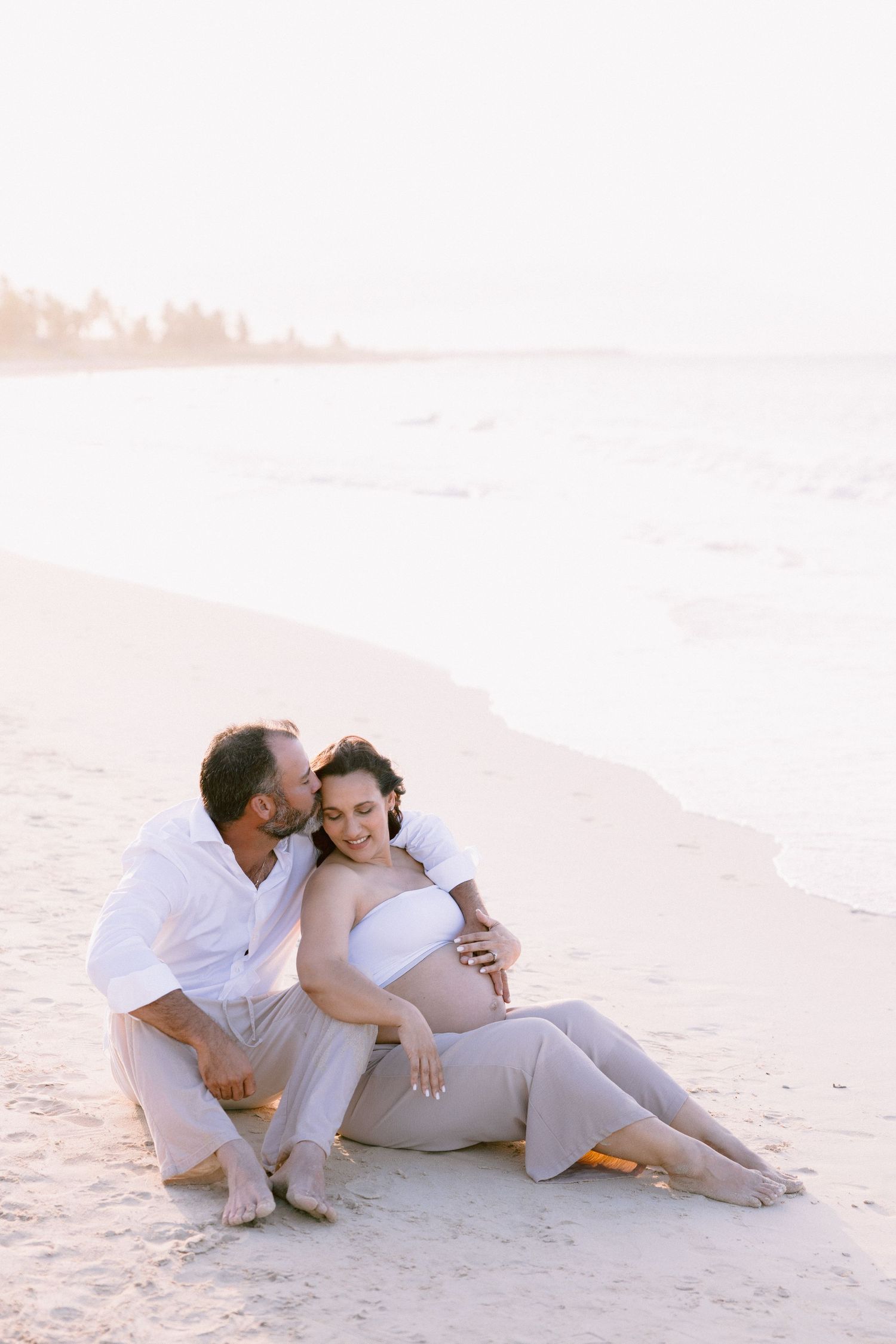 A couple sits on a sandy beach at sunset, man in white shirt embracing pregnant woman in white top and gray skirt.