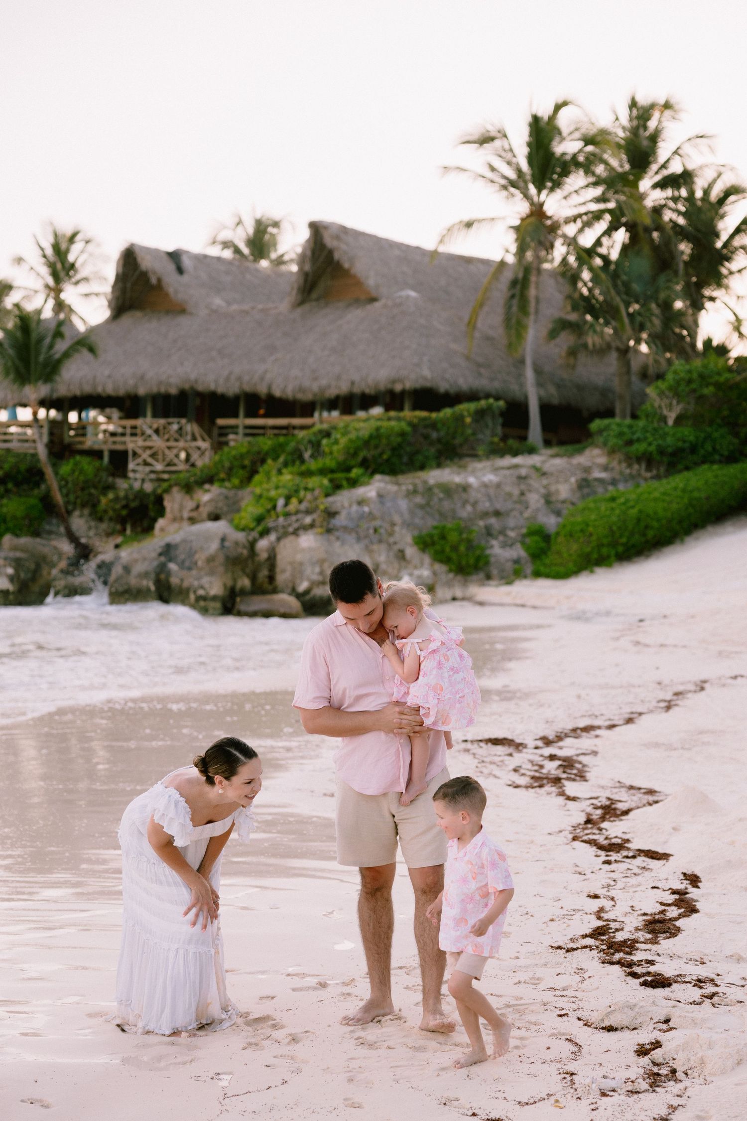 Family of four enjoying a tropical beach vacation at sunset near a thatched-roof resort with palm trees.