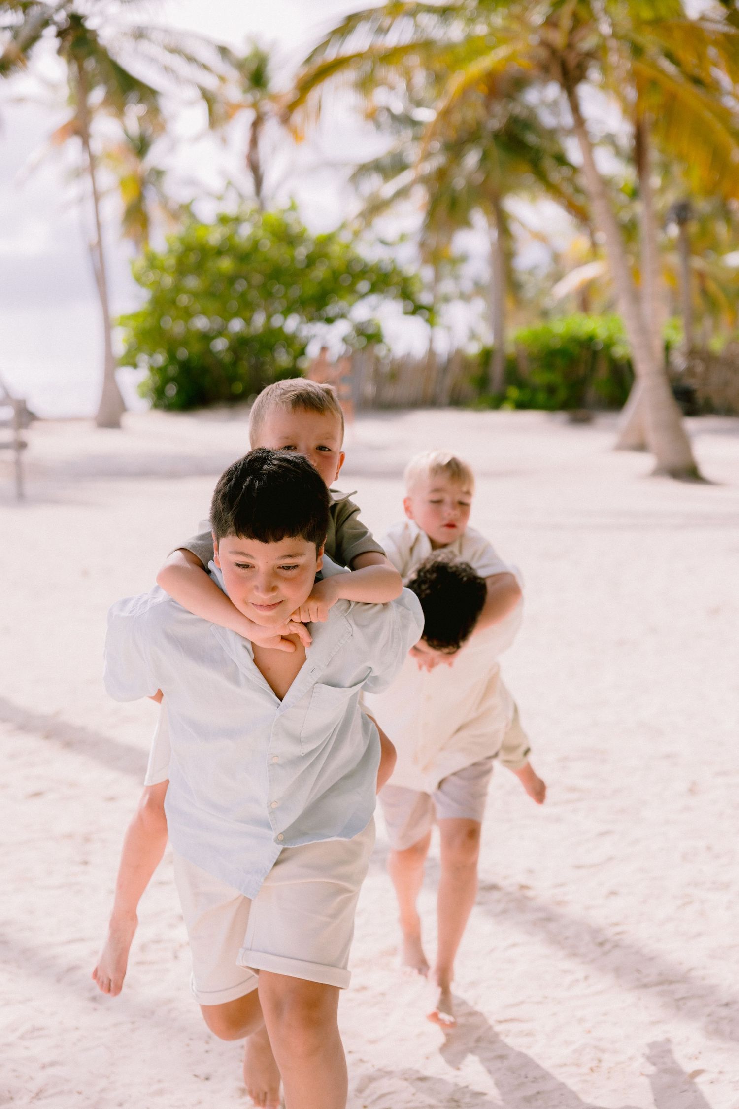 Children playing piggyback rides on a sandy tropical beach surrounded by lush palm trees on a sunny day.