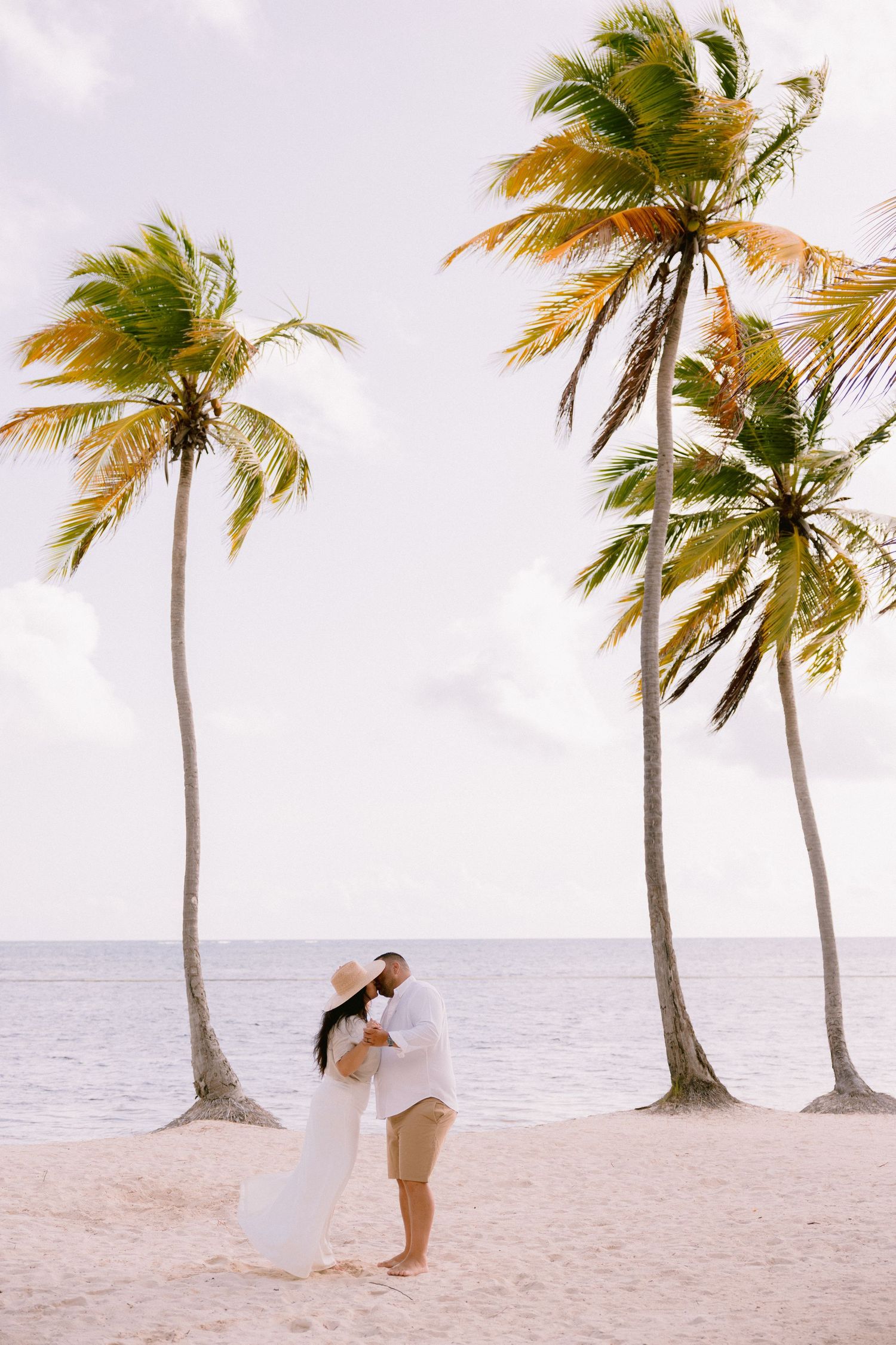 Couple kissing on a tropical beach surrounded by tall palm trees with ocean views on a sunny day.