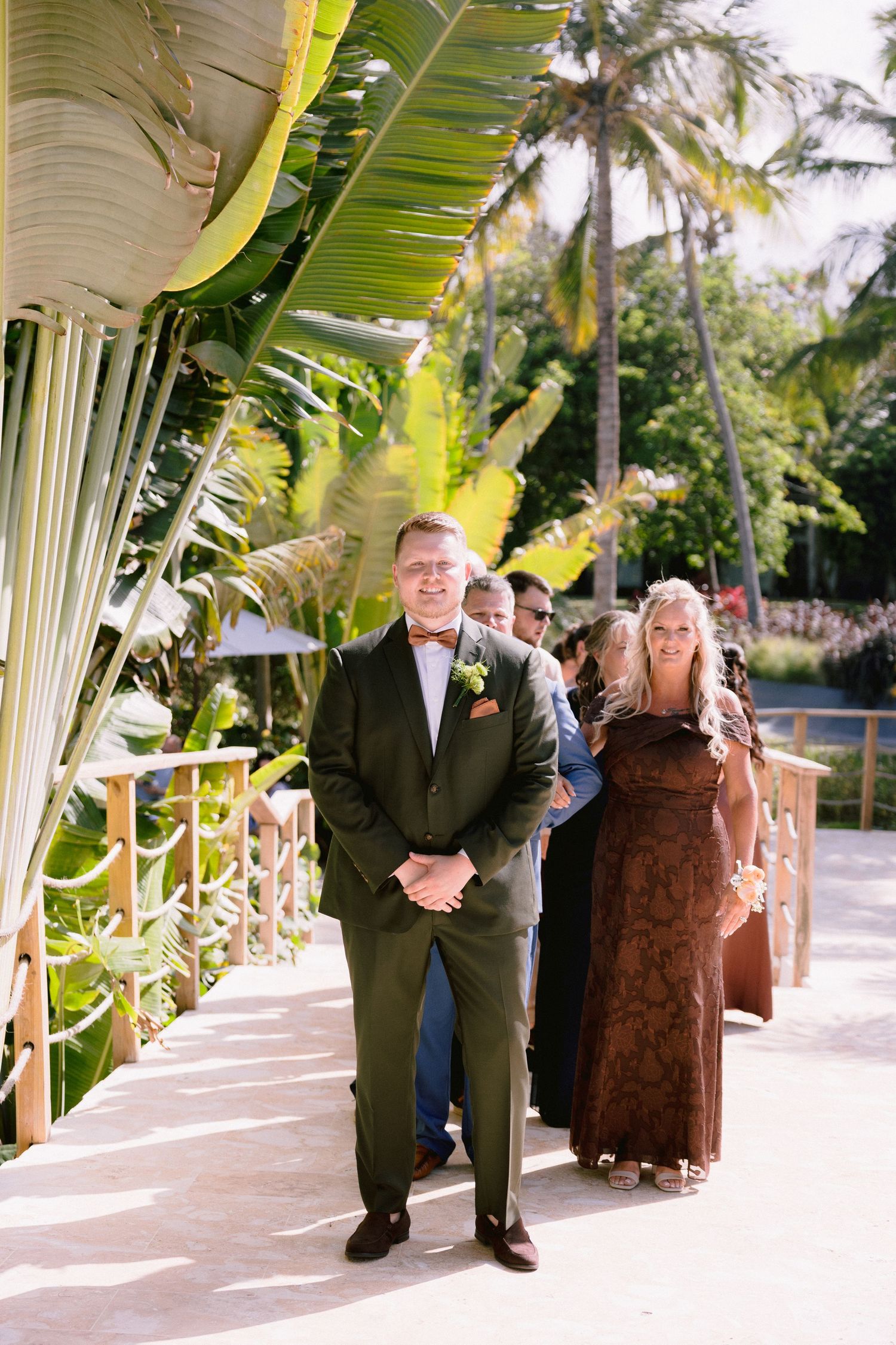 Groom in olive suit with boutonniere walks down tropical outdoor aisle with guests and palm trees at wedding.