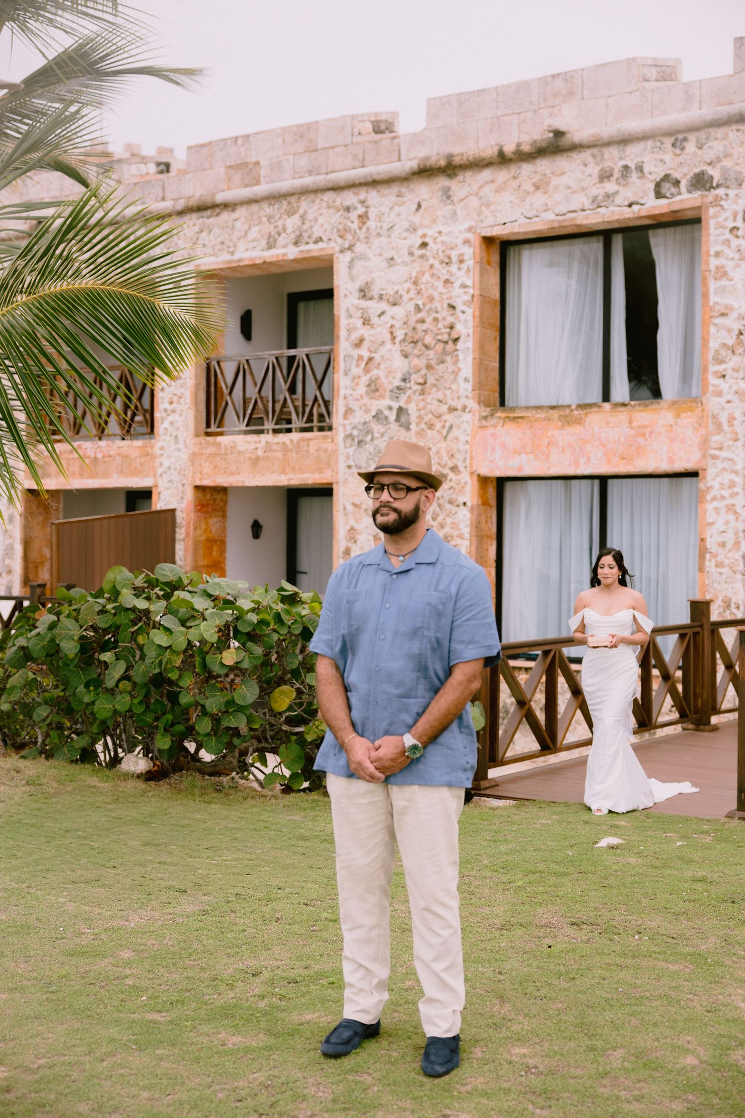 Groom in blue shirt and hat stands outdoors at resort while bride in white gown approaches from behind.