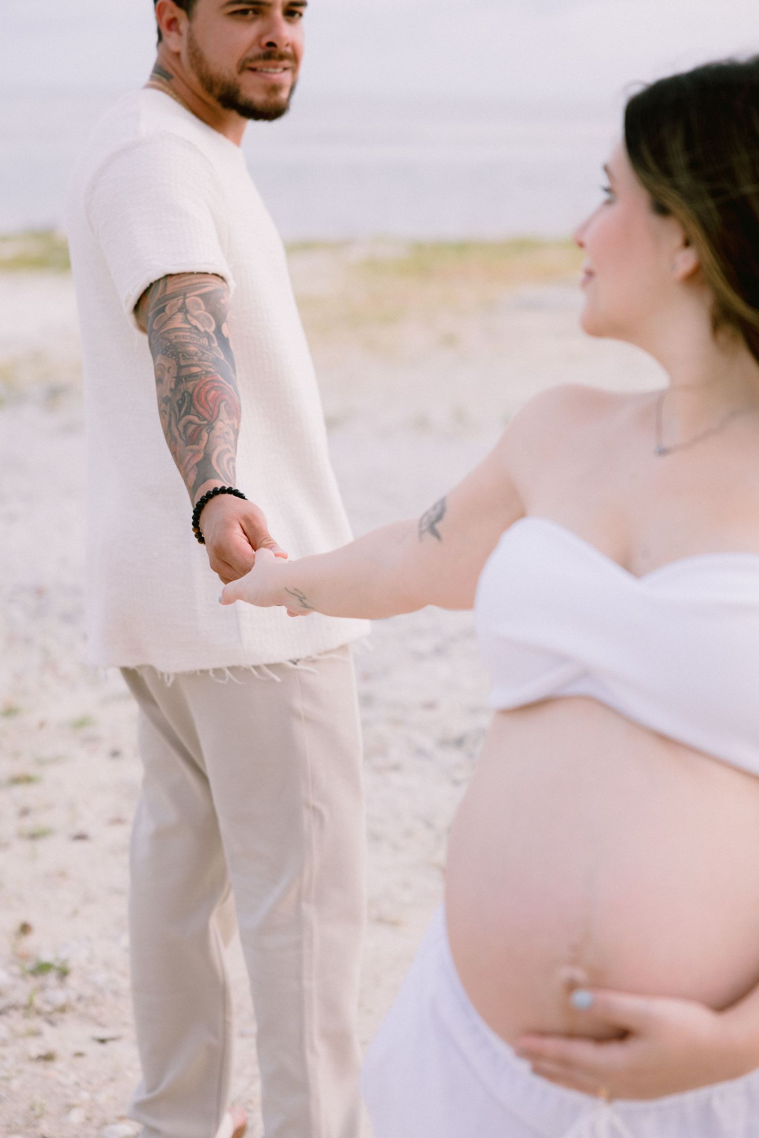 A couple in white wedding attire embraces lovingly on a beach during a romantic photoshoot.