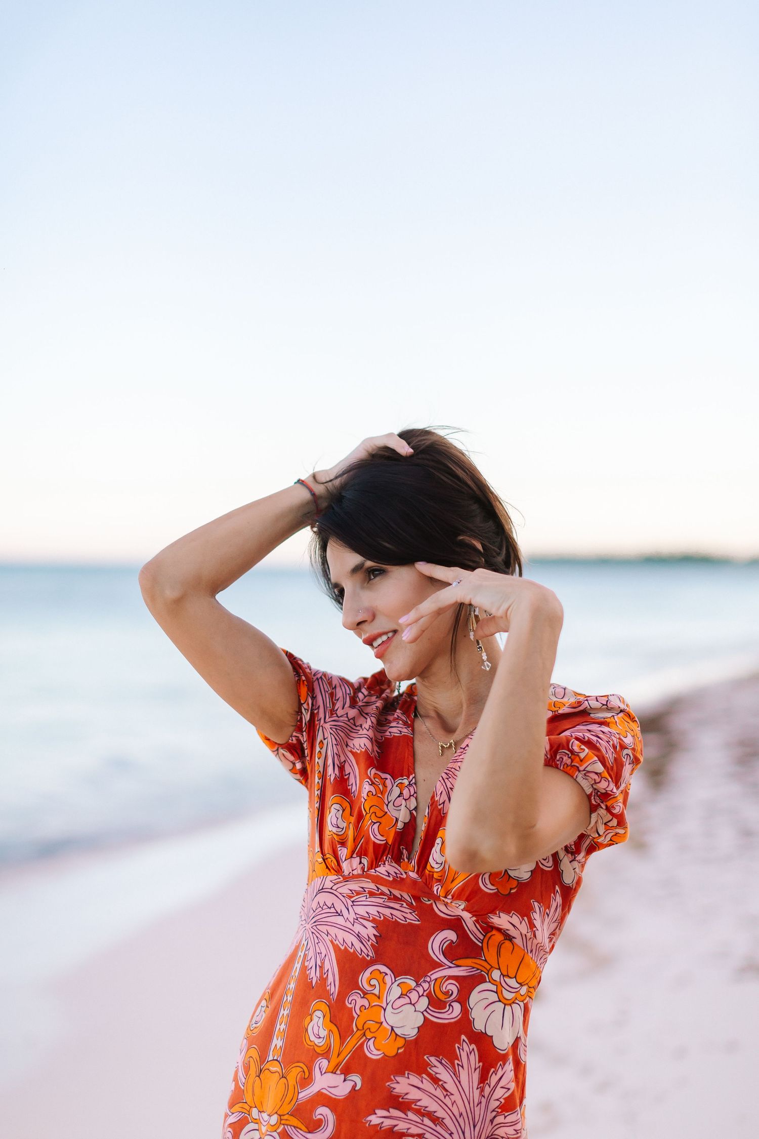 A woman in a vibrant orange floral dress poses on a beach during golden hour.