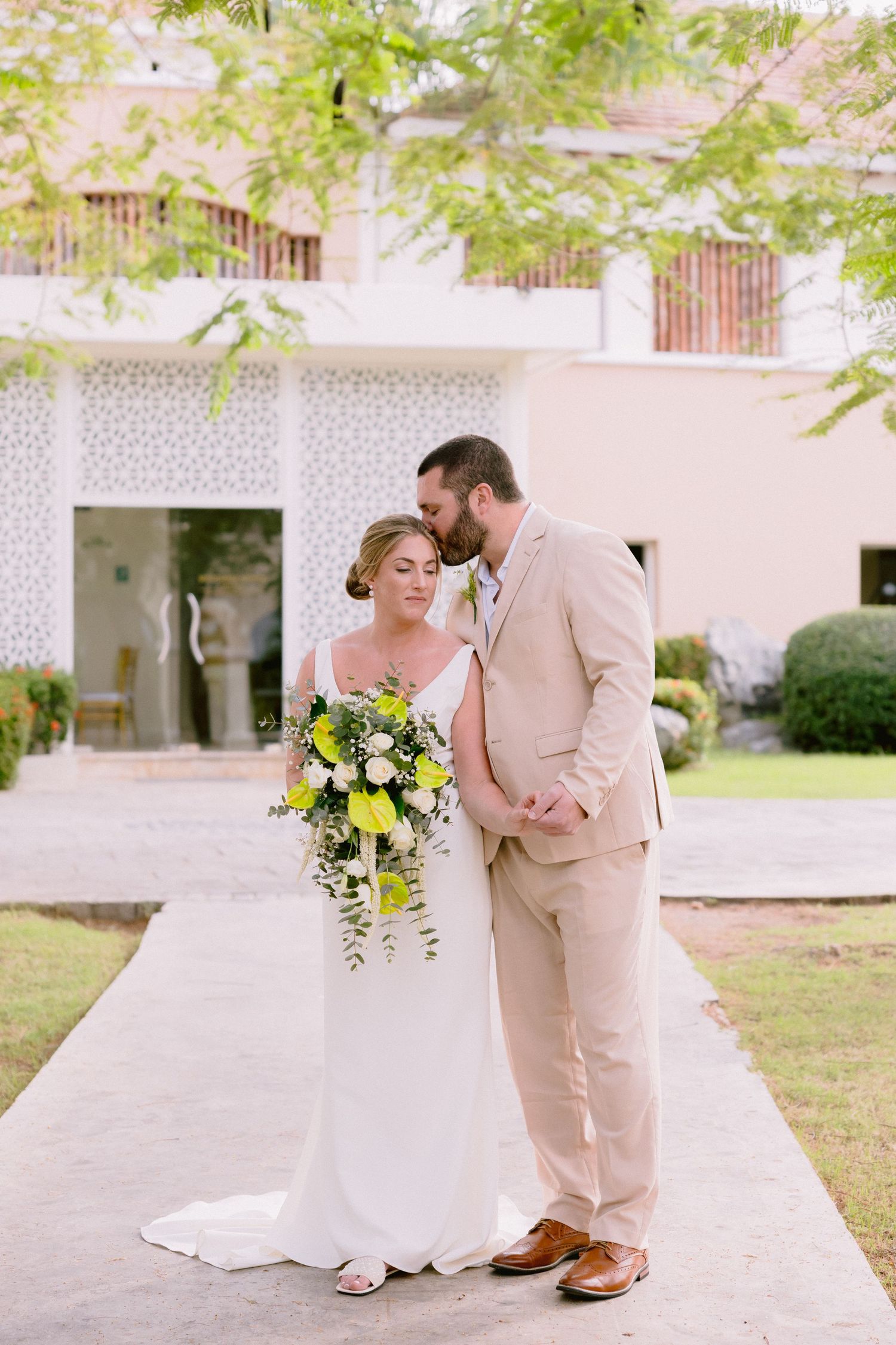 Bride and groom share a tender moment outdoors, she holds a green and white bouquet, he wears a beige suit.