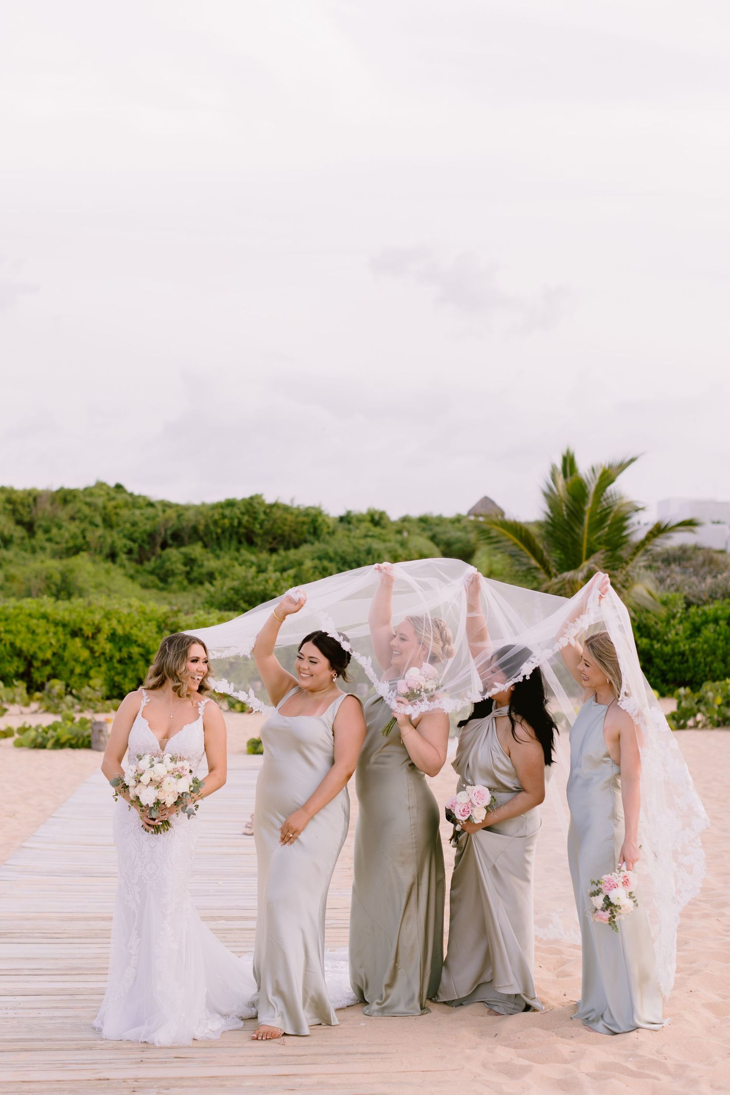 Bride and bridesmaids in sage green gowns laughing as veil blows in wind on sandy beach boardwalk.