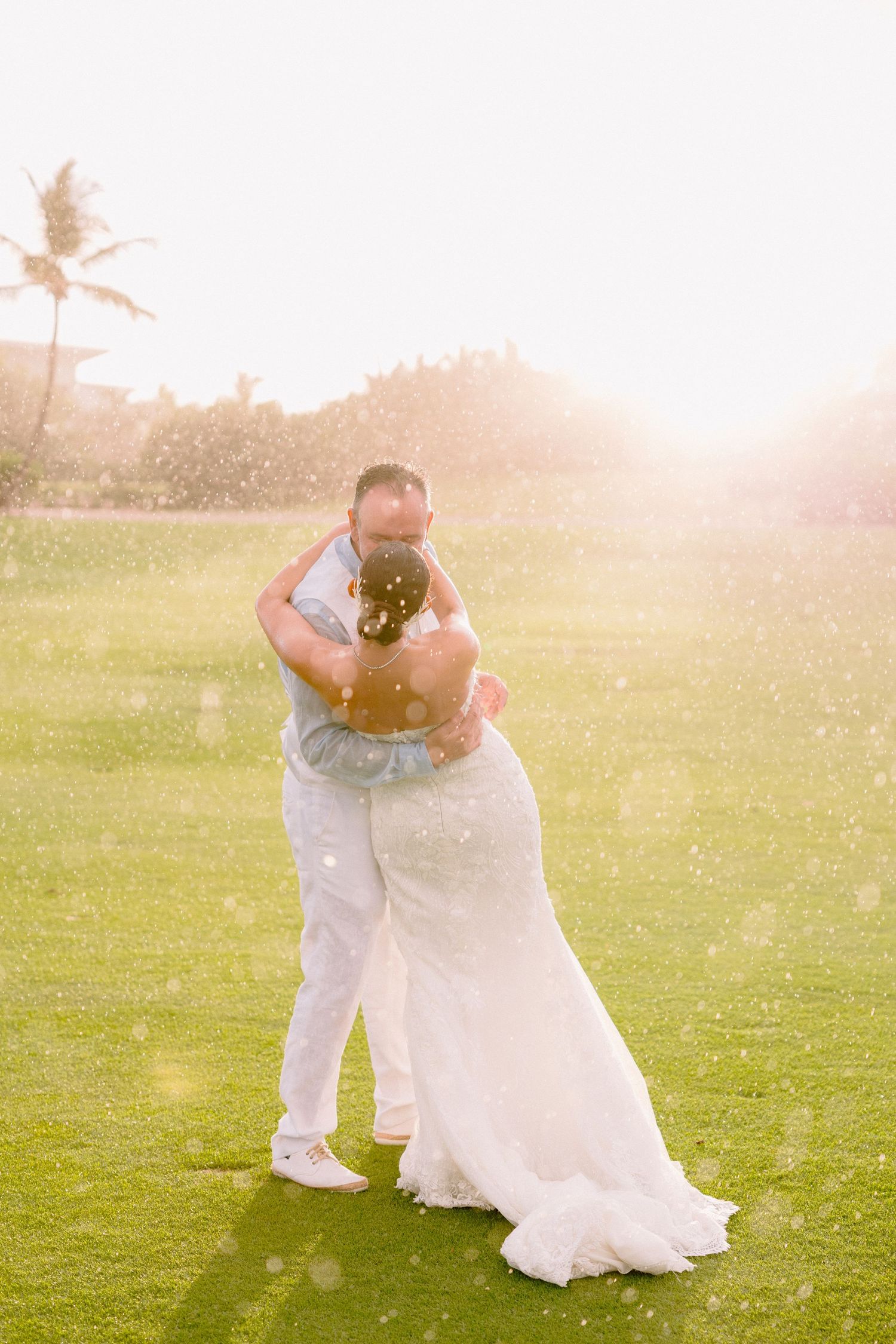 A couple in white attire embraces during a sunset photoshoot on a grassy field with golden sunlight streaming through.