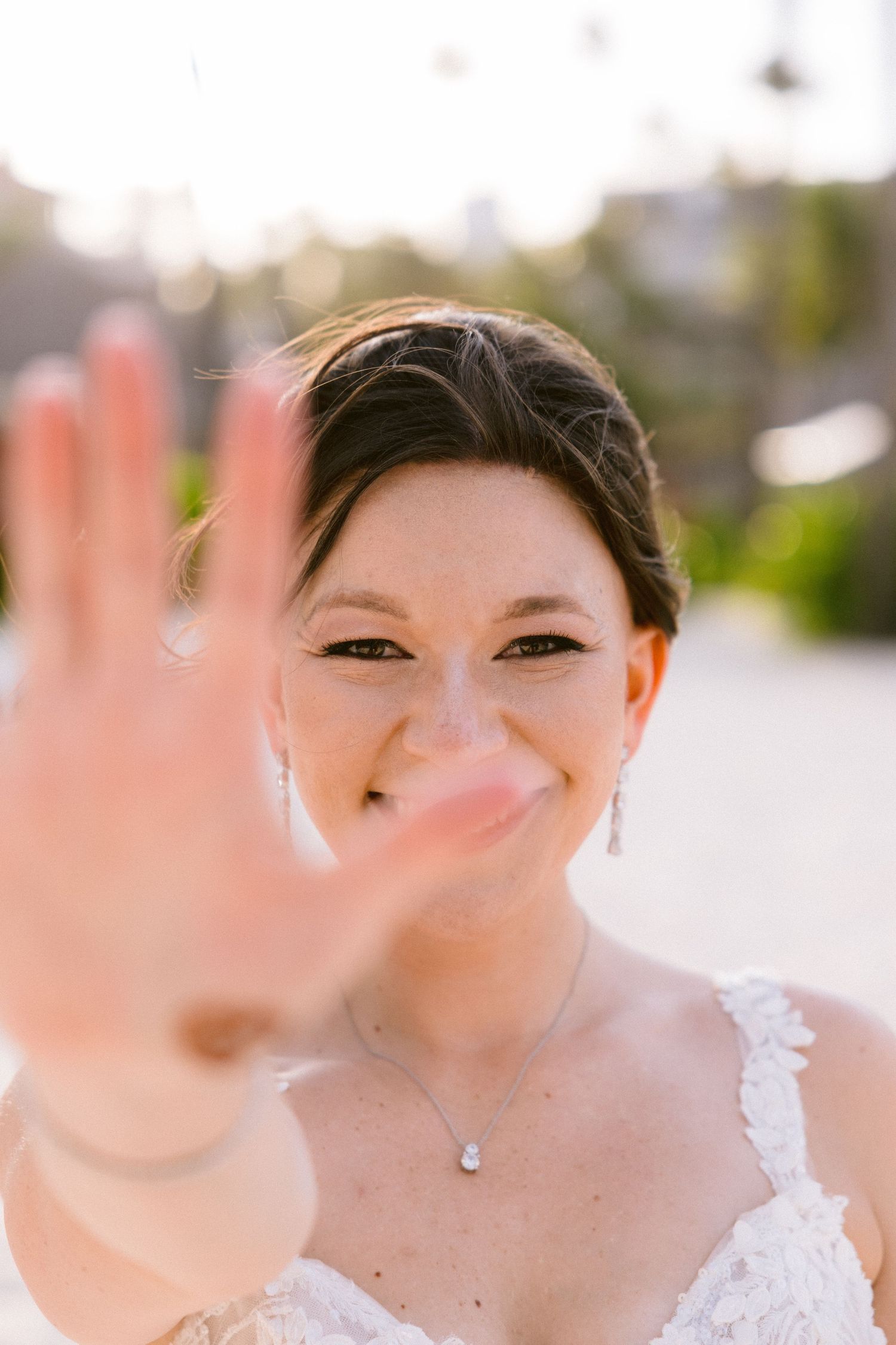 A bride in a white lace dress smiles and holds up her hand to the camera on her wedding day.