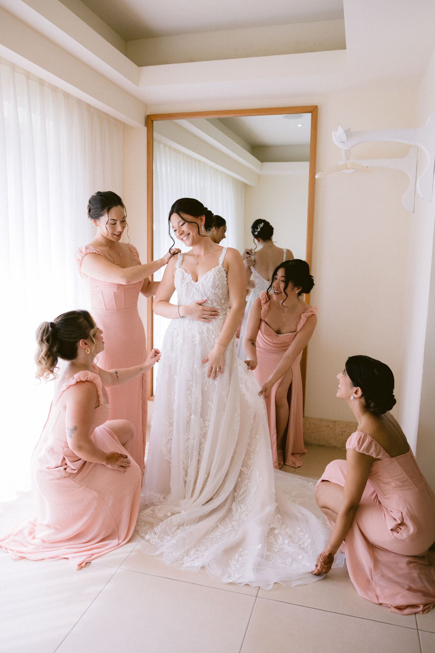 Bridesmaids in blush pink dresses help the bride get ready in a sunlit room with a white gown and soft decor.