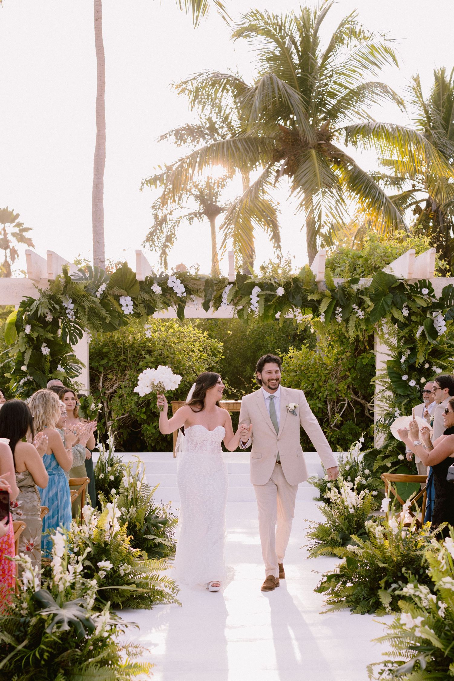 Wedding guests gather on a white beach walkway surrounded by tropical palm trees and lush greenery at sunset.