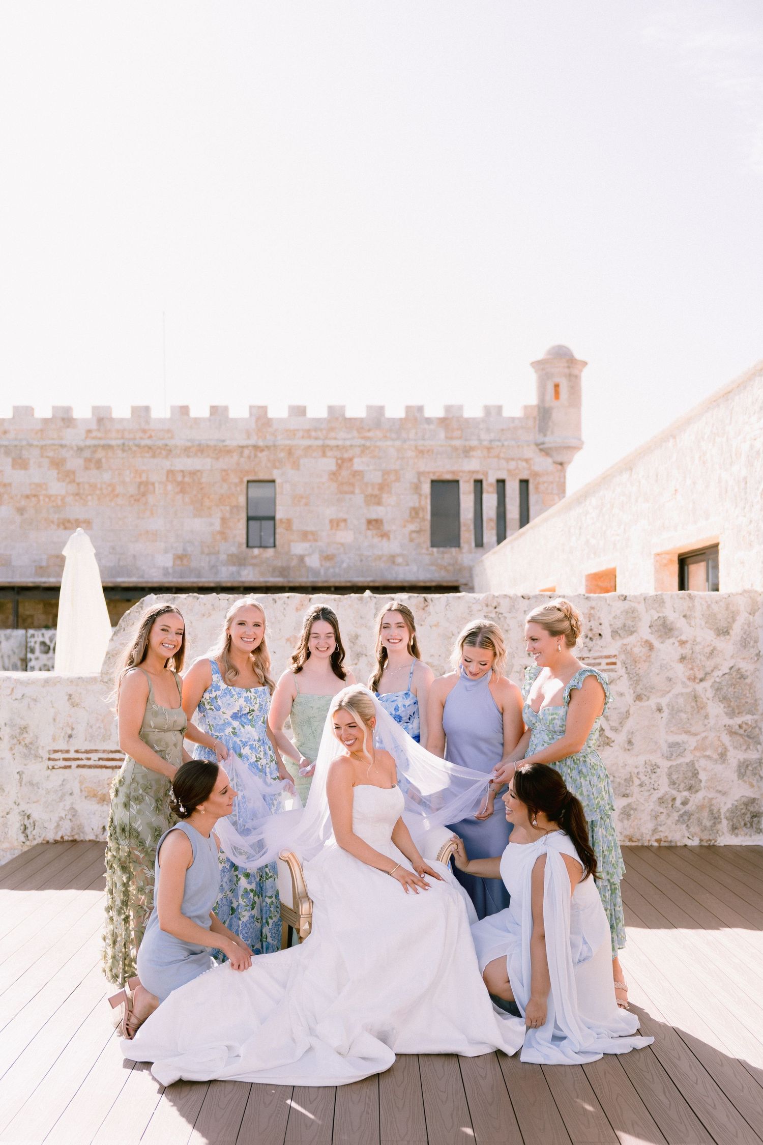 Bride in white gown seated with bridesmaids in blue and floral dresses on a sunny rooftop terrace.