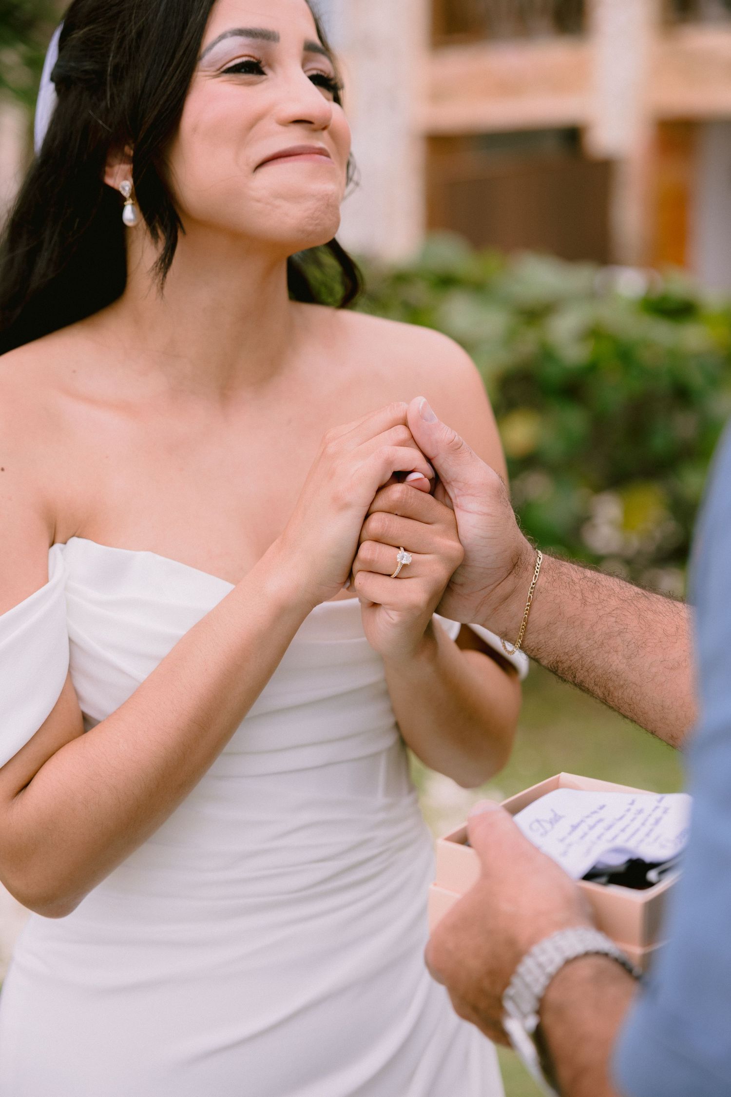 Emotional bride in white strapless gown holds groom's hand during outdoor wedding ceremony vow exchange.