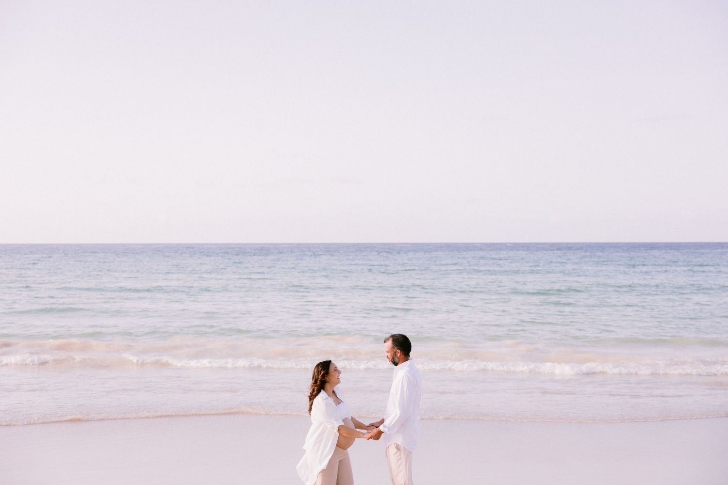Couple in white outfits holding hands on a serene beach with turquoise ocean waves in the background.