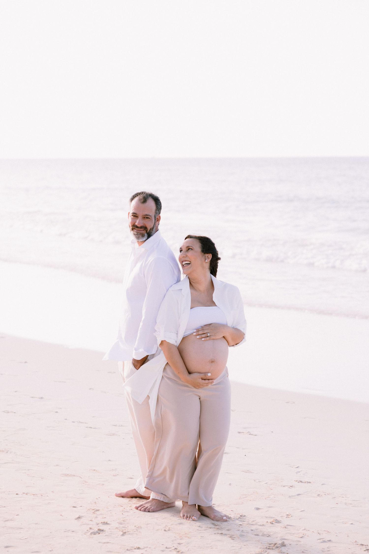 Happy pregnant couple standing barefoot on a serene beach, woman cradling her baby bump while leaning against her smiling partner.