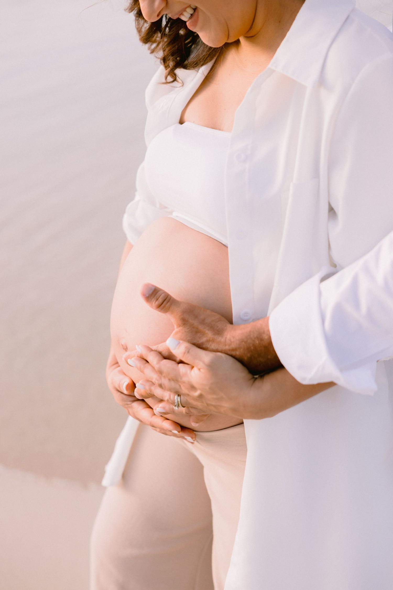 Pregnant woman in white top and sheer shirt with partner's hands cradling baby bump on a serene beach.