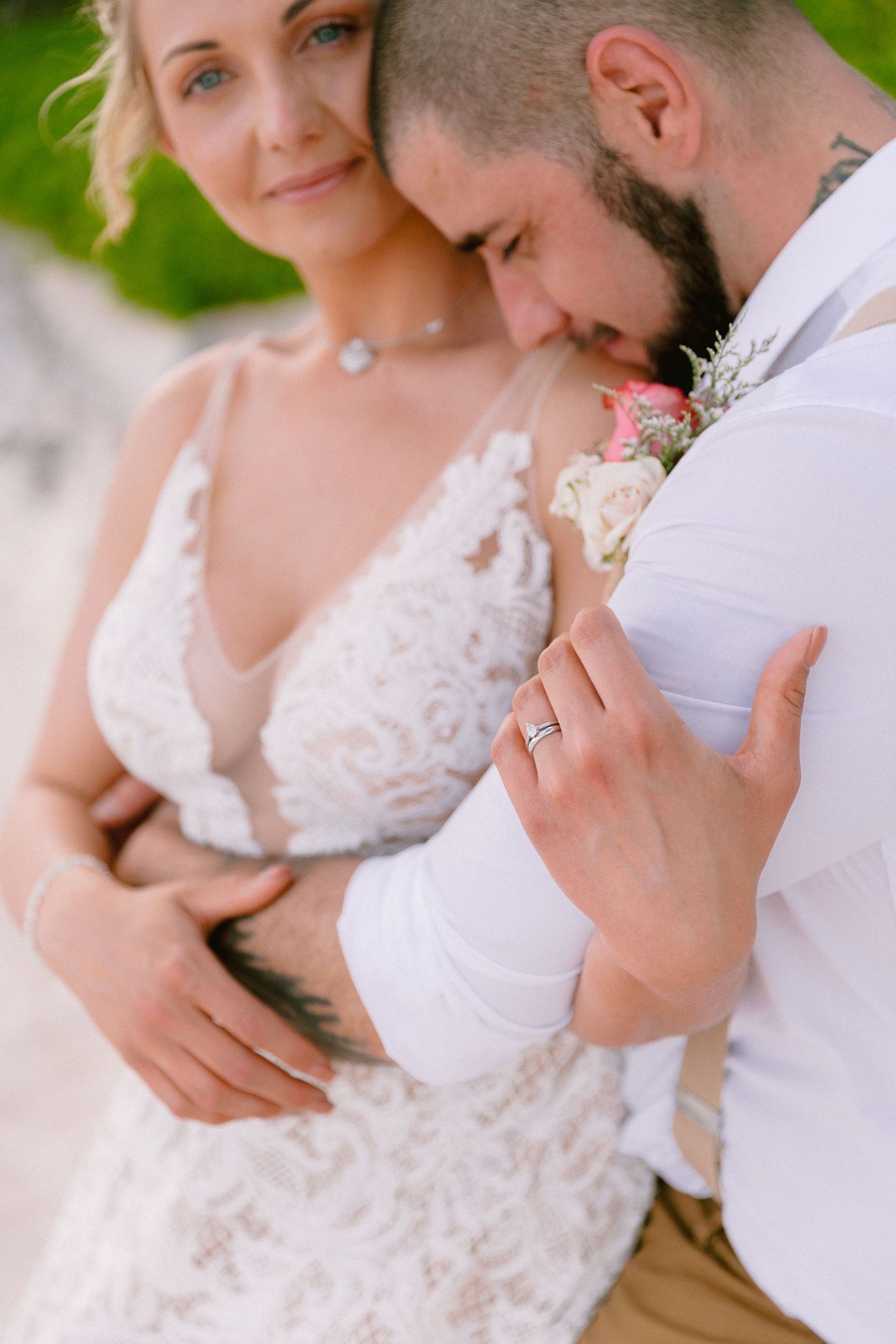 Bride in lace gown embraces groom in white shirt, showcasing wedding rings during romantic outdoor ceremony.