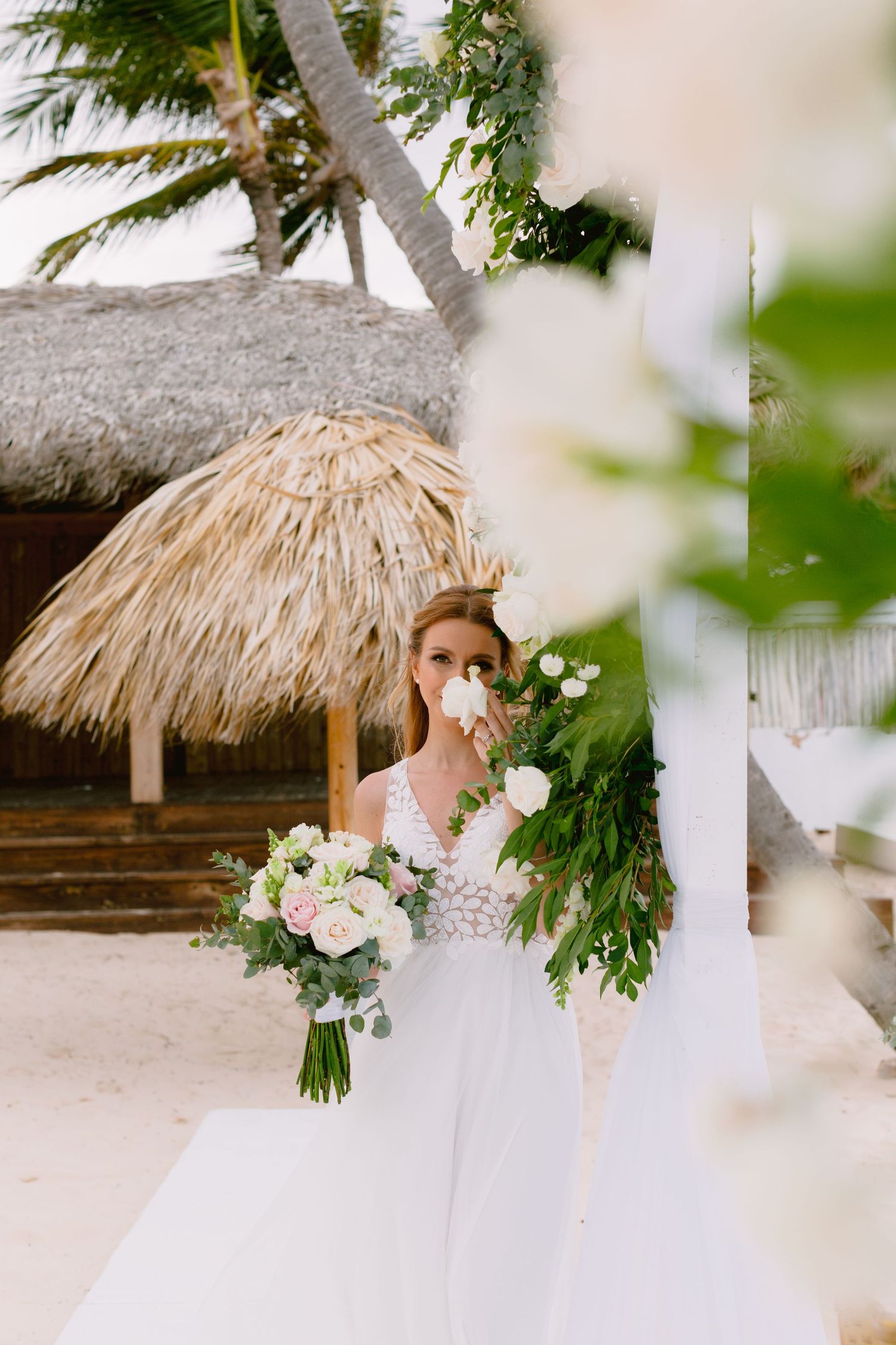 Bride in white lace gown holding pink and white bouquet at tropical beach wedding ceremony with palm trees.