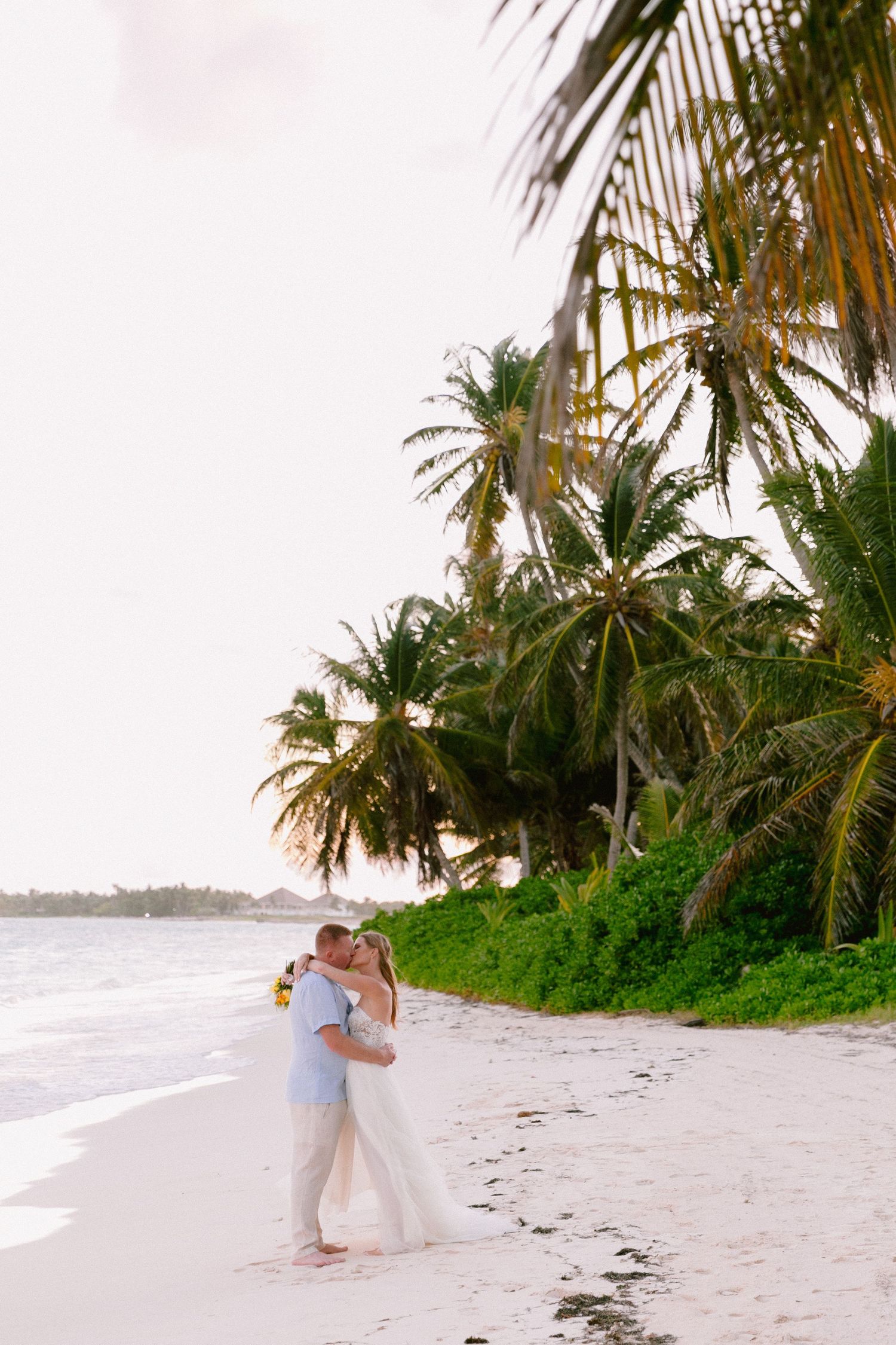 Couple kissing on a tropical beach with palm trees and white sand at sunset.