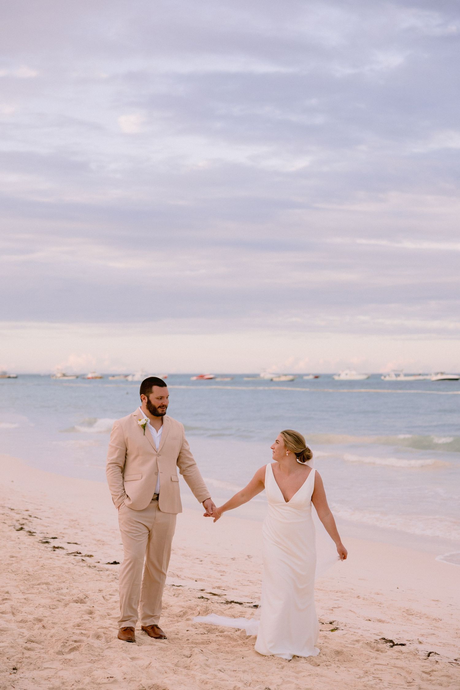 Bride and groom hold hands walking on a sandy beach at sunset, with calm turquoise water and boats in the background.