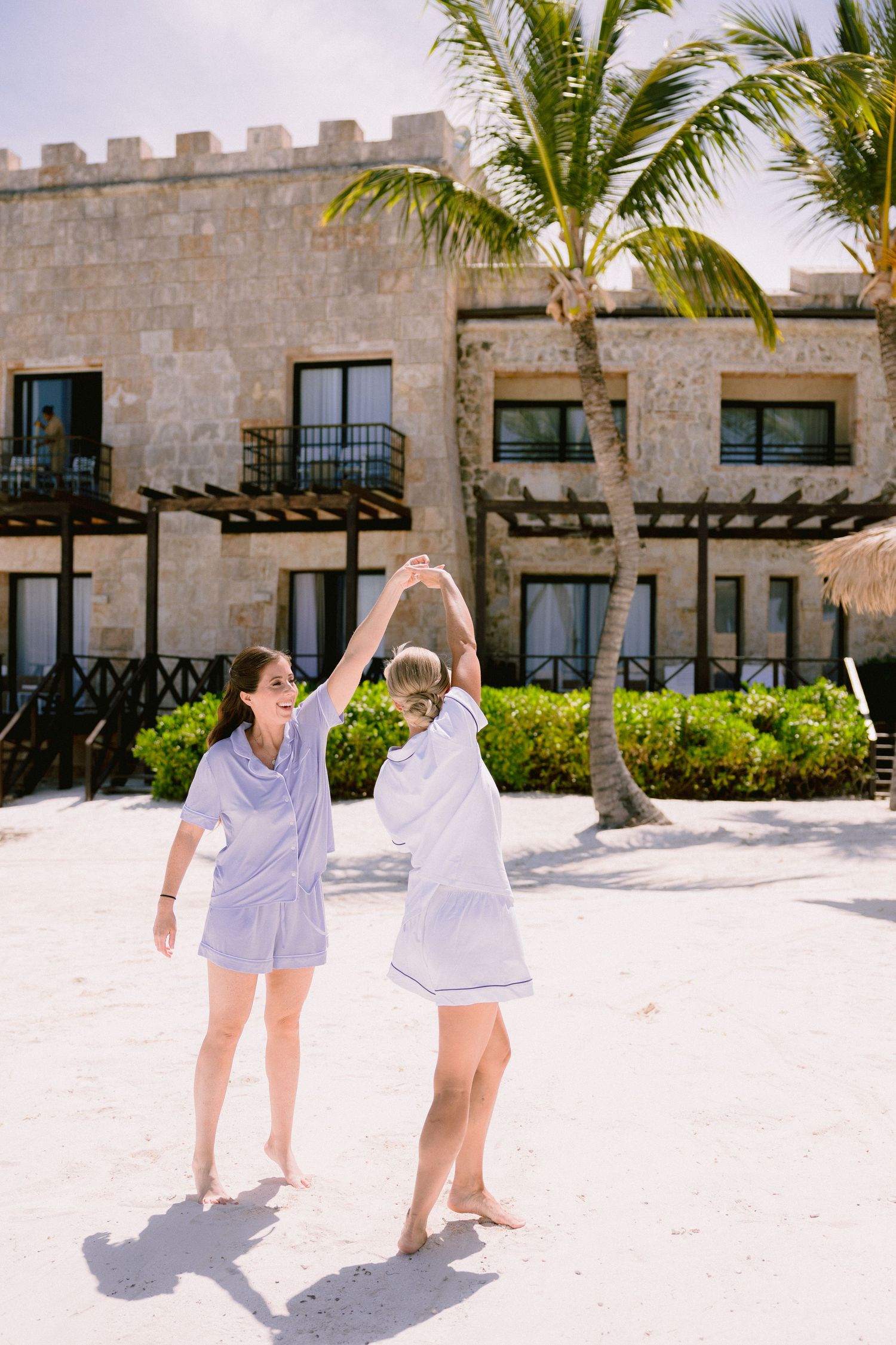 Two women in matching blue outfits dancing on white sand beach near palm trees and a stone resort building.