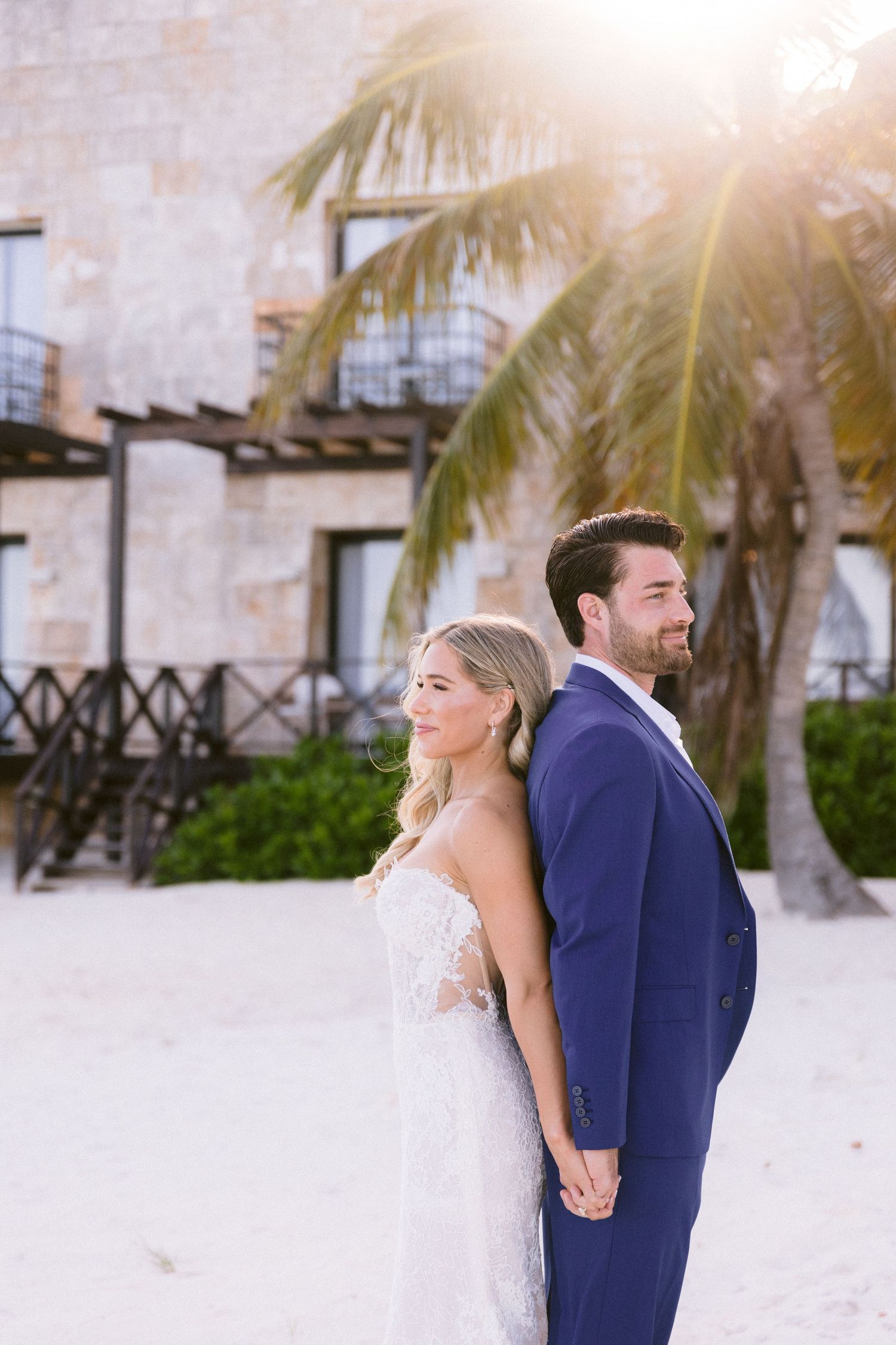 Bride and groom stand back-to-back on a sunny beach, wearing a lace gown and navy suit, with a palm tree behind them.