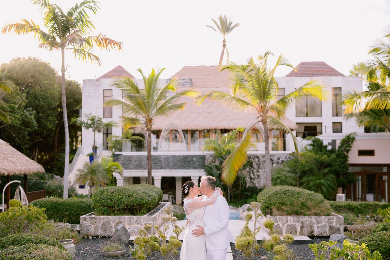 A beautiful wedding ceremony takes place at sunset in a tropical garden setting with palm trees and elegant decor.