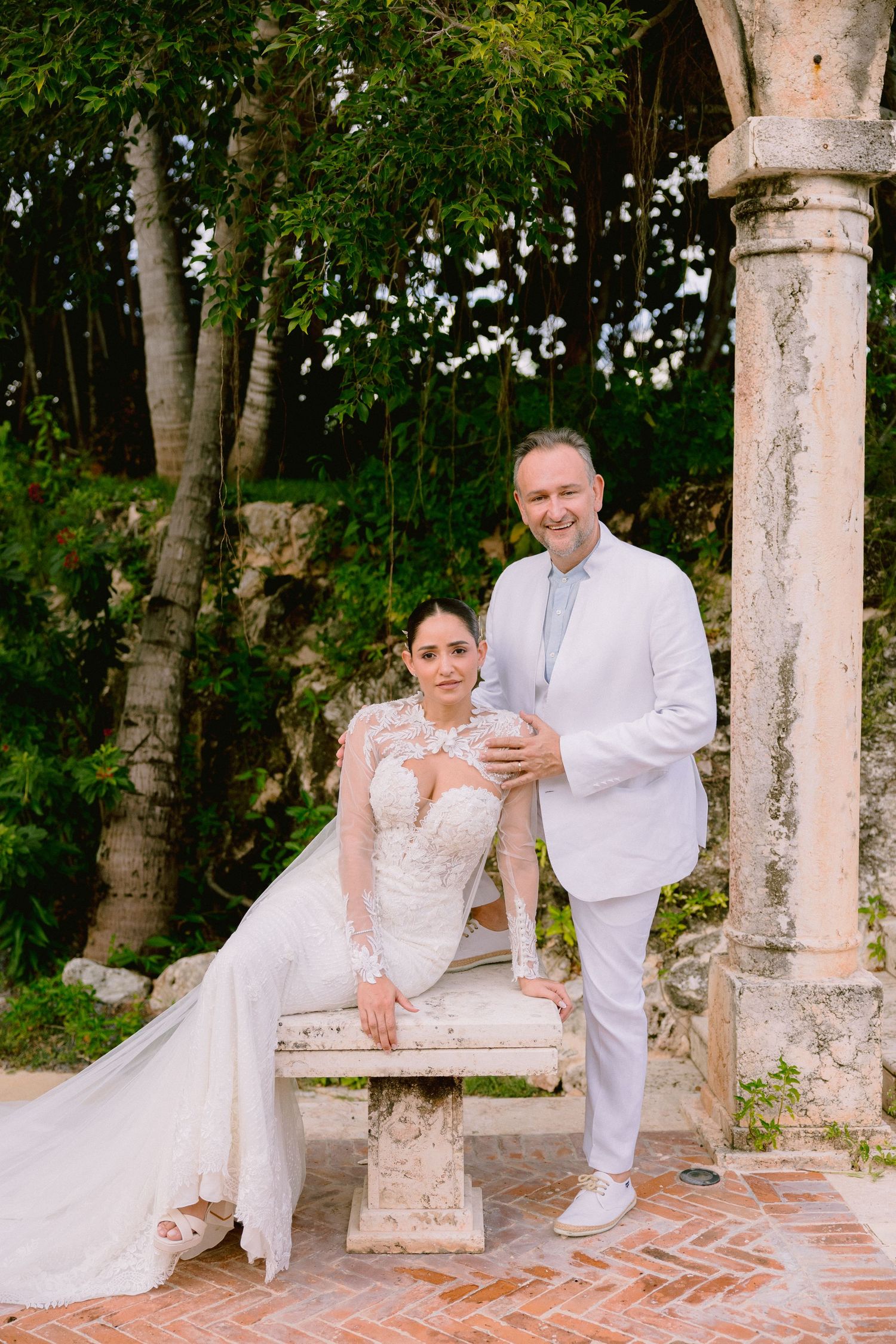 A wedding couple poses together on stone columns and brick steps in a lush garden setting.
