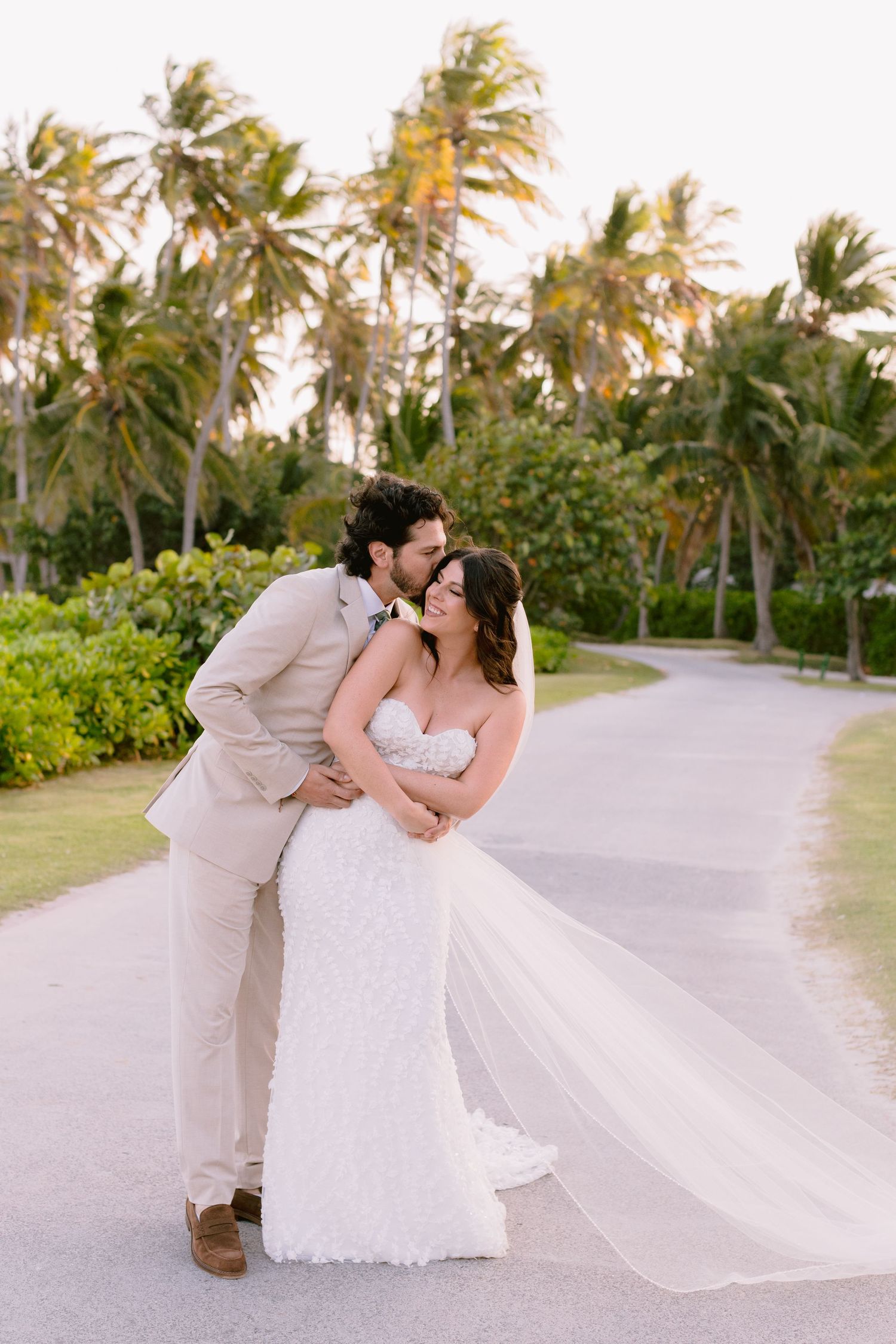 Romantic wedding couple embraces on a palm-lined tropical path during sunset.