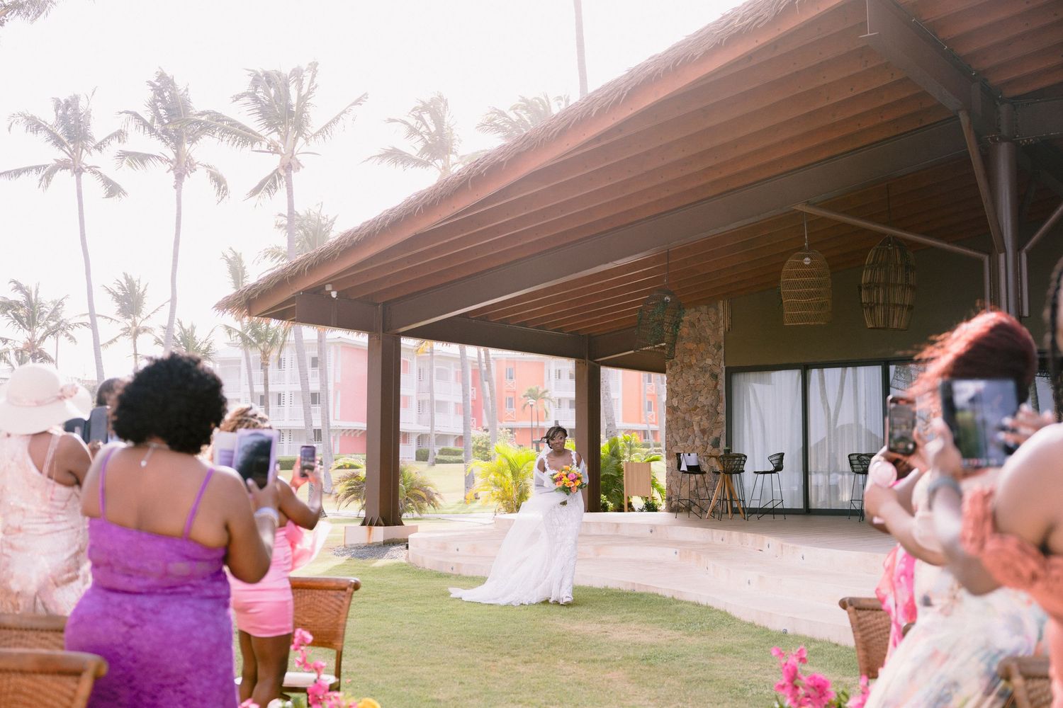 Outdoor party scene with people gathered under a covered patio area near palm trees on a sunny day.