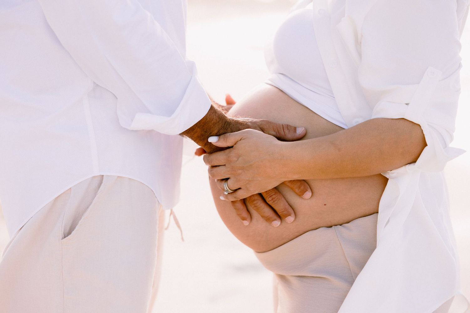Two people in white clothing holding hands over a pregnant belly outdoors.