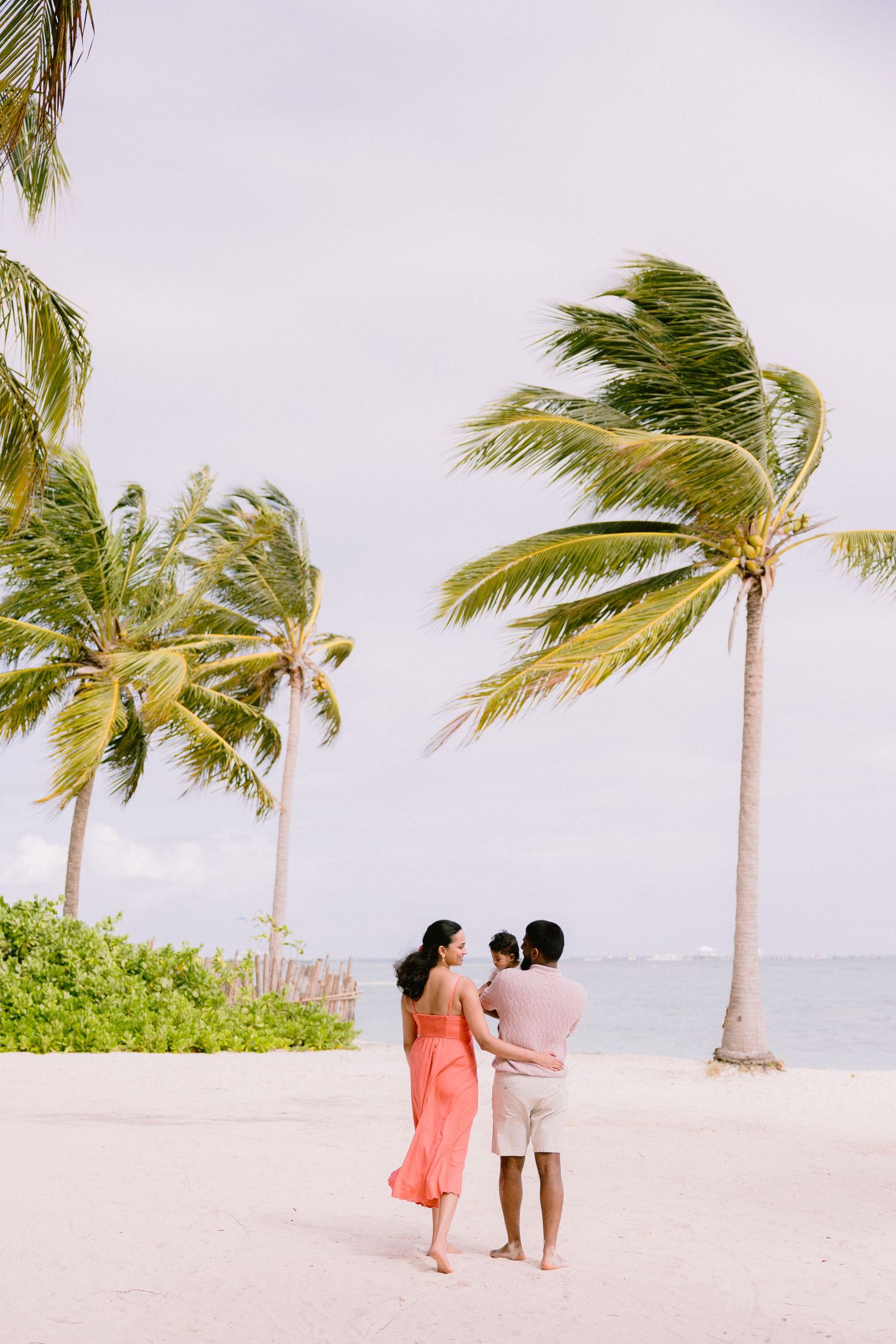 Couple with baby on a tropical white sand beach with palm trees swaying in the breeze under a cloudy sky.