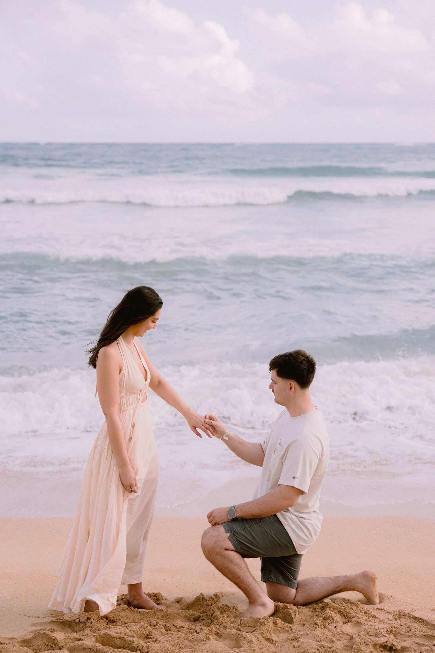 Man proposes to woman on sandy beach with ocean waves in background.