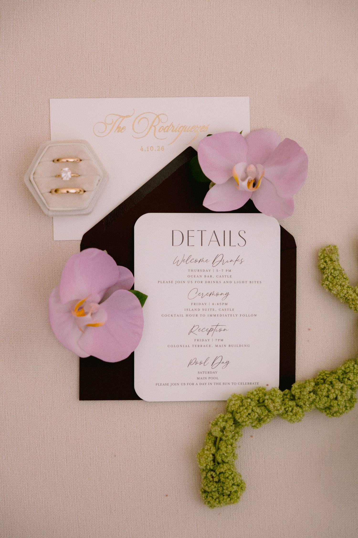Elegant wedding invitation suite with details card, black envelope, pink orchids, and diamond ring in velvet box on neutral background.