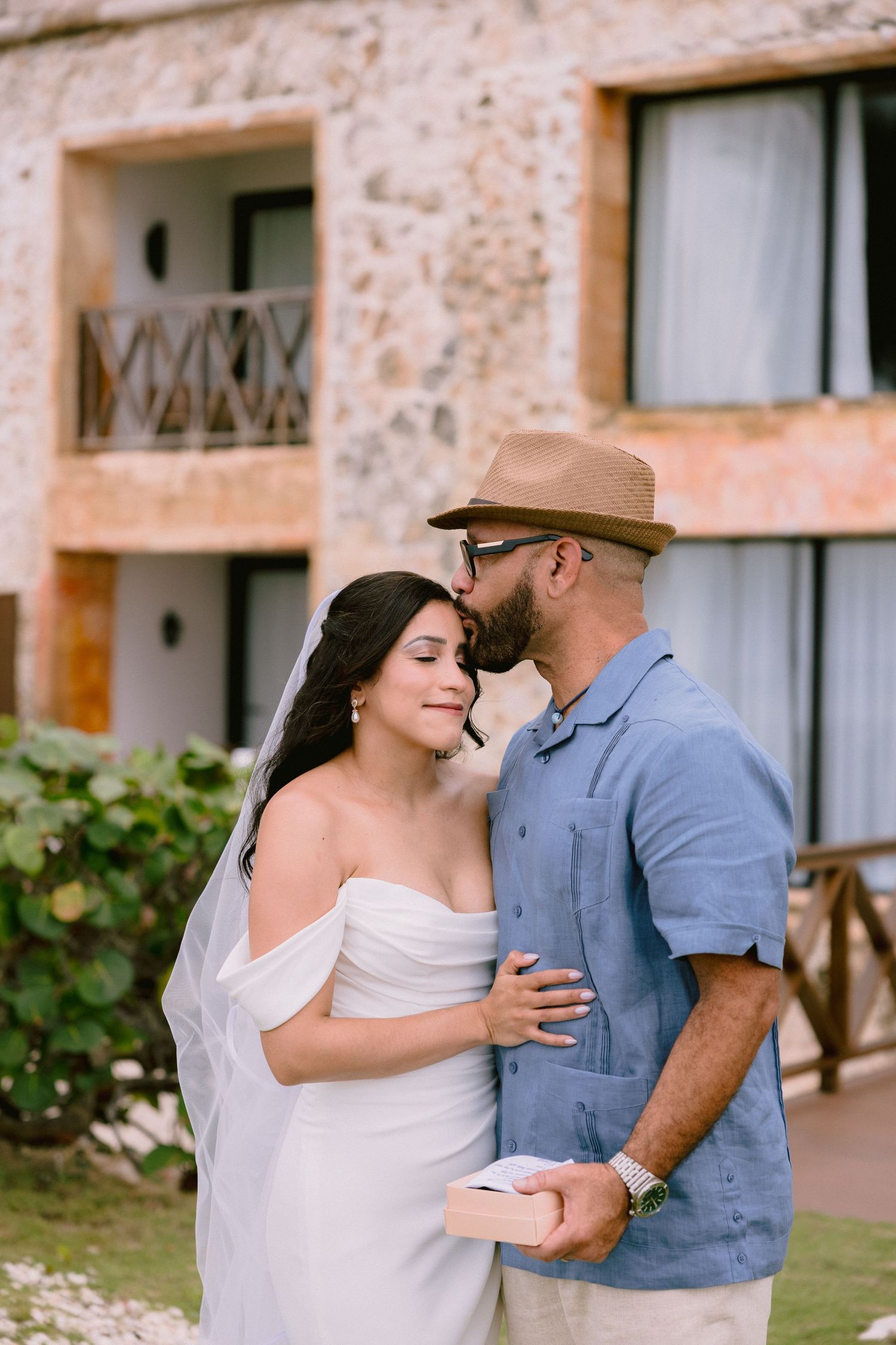 Bride in white off-shoulder gown and veil embraced by groom in blue shirt and tan hat at a resort.