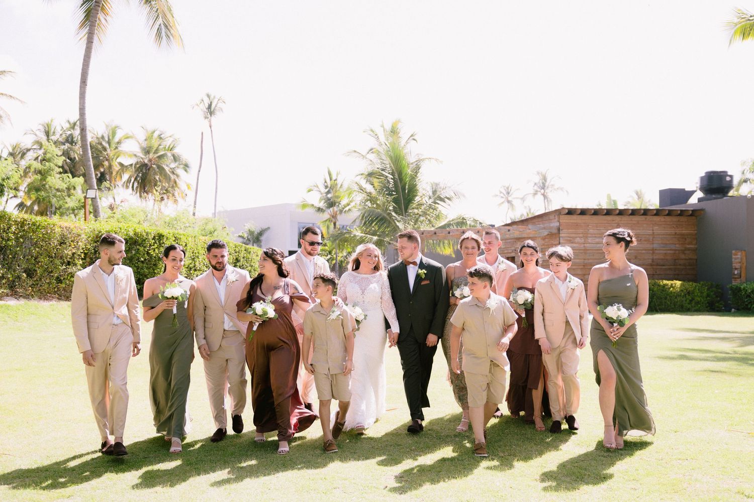 Wedding party walks outdoors on green lawn with palm trees, bride in white gown surrounded by groomsmen and bridesmaids.