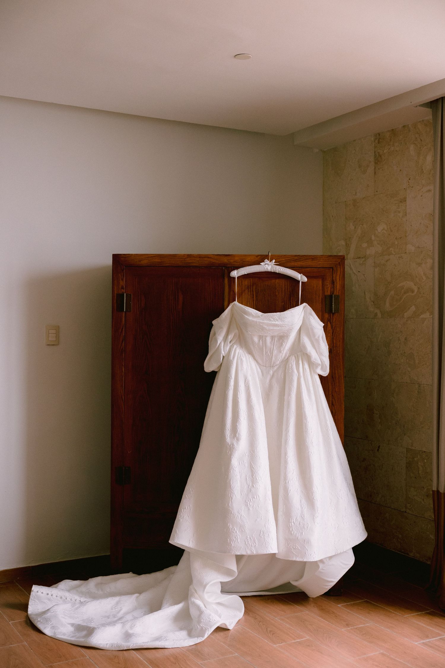 White off-shoulder wedding dress with long train hanging on a wooden wardrobe in a softly lit room.