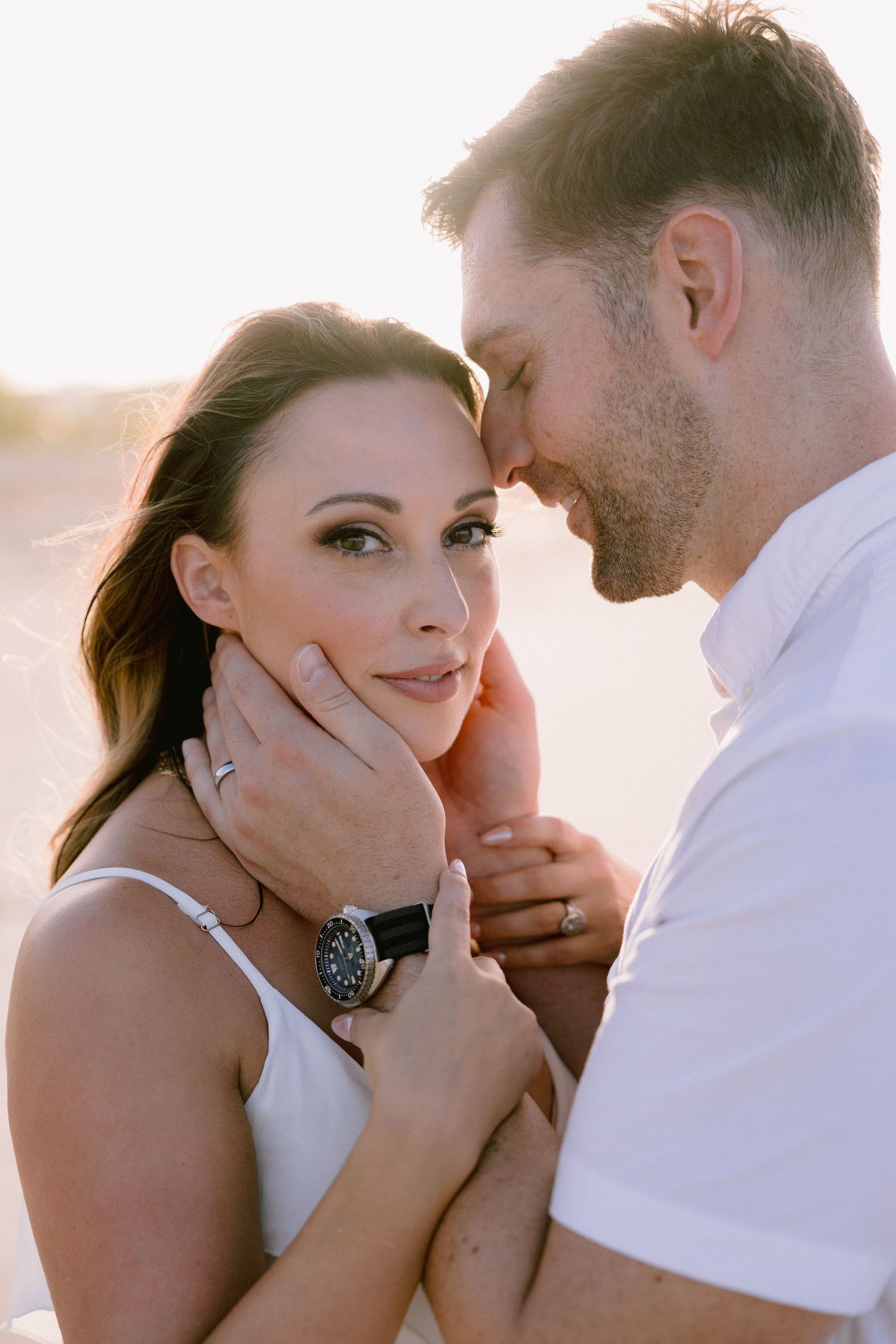 Romantic couple embracing at sunset on a beach, woman in white dress with engagement ring, man tenderly holding her face.