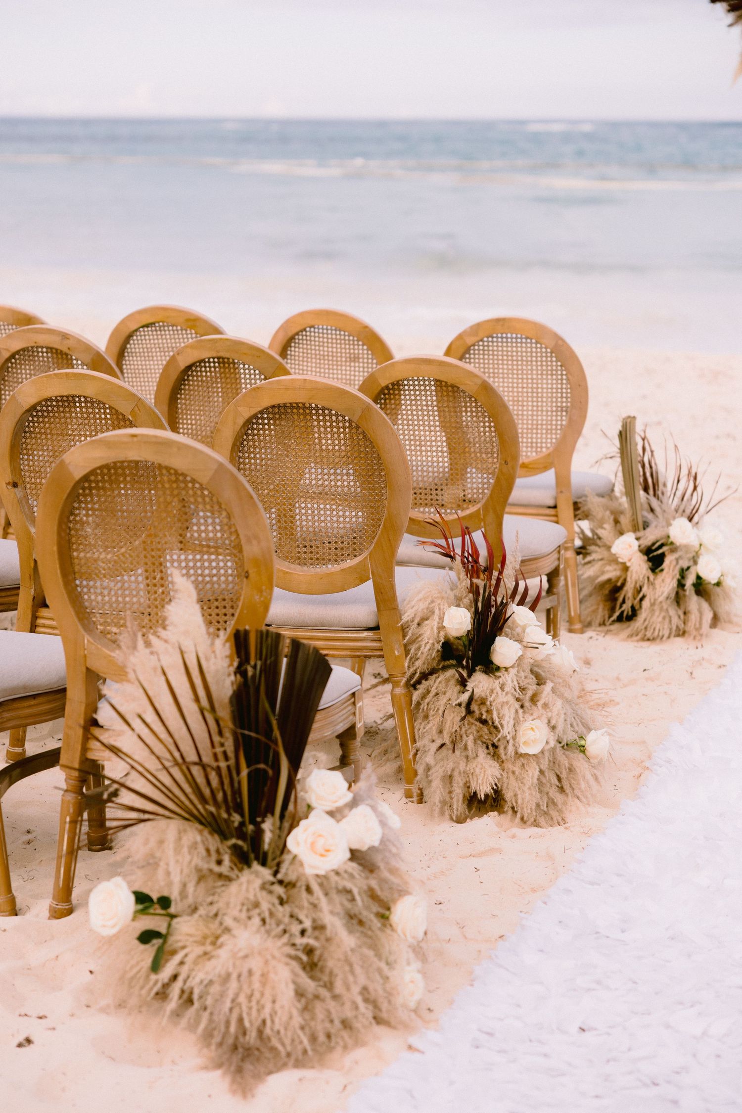 Elegant rattan chairs arranged on white sand beach for a boho wedding ceremony with pampas grass floral arrangements.